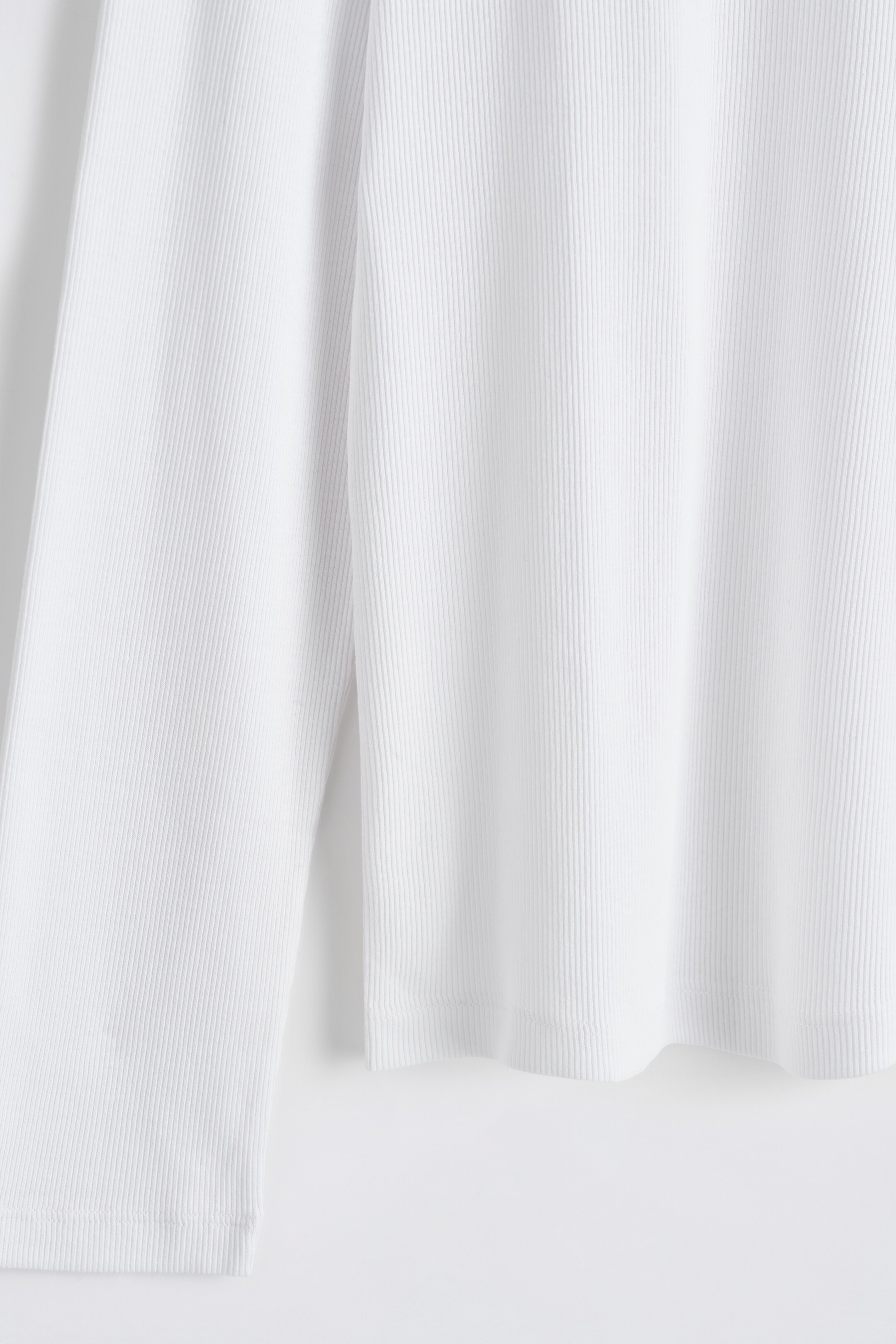 Visualizza immagine più grande: Un top bianco a maniche lunghe è mostrato dalla parte anteriore, caratterizzato da una texture finemente a coste che corre verticalmente lungo il tessuto. Il suo orlo inferiore dritto e il polsino delle maniche sono visibili, presentando un design aderente.
