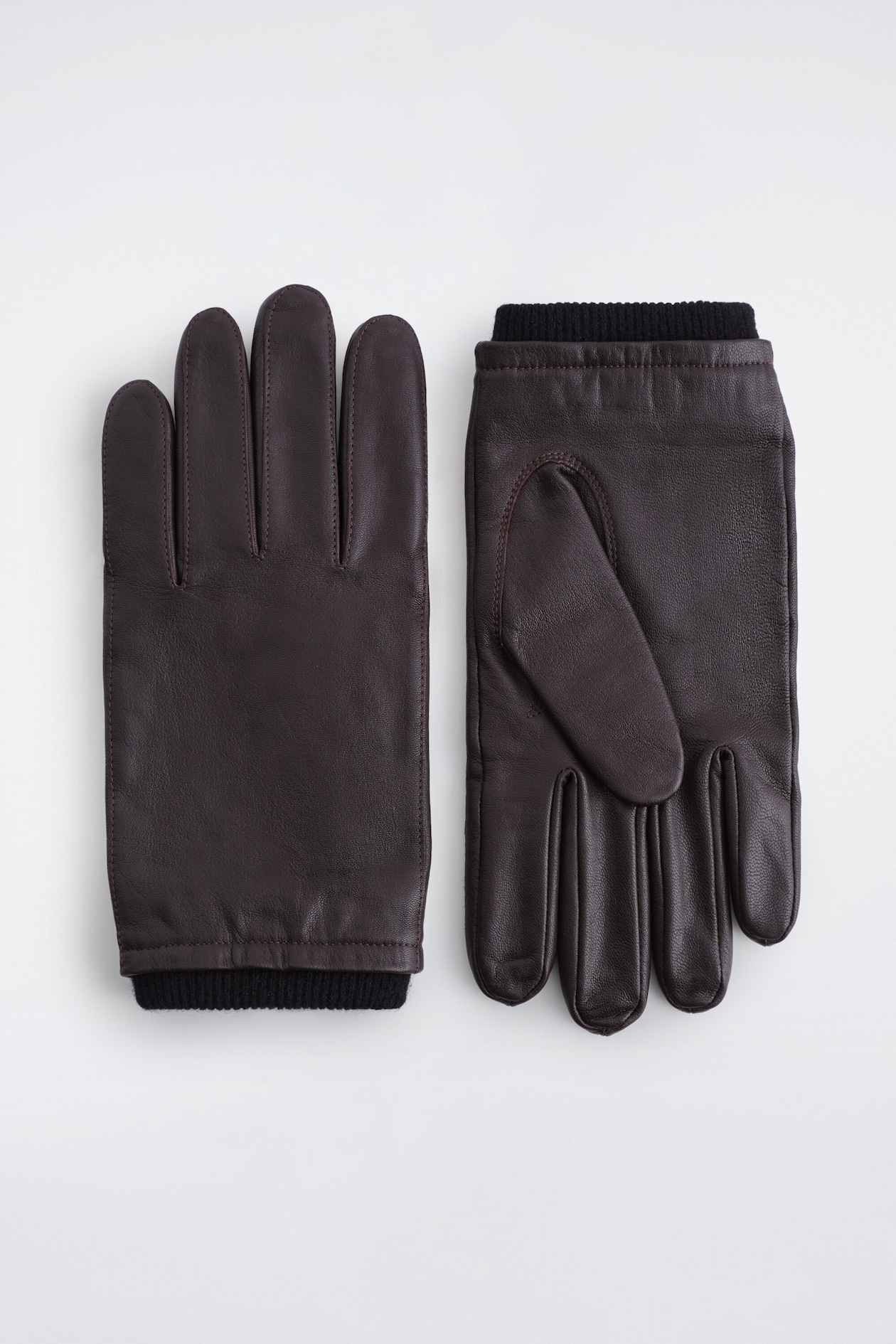 Topstitched leather gloves in dark brown are laid flat, with the left glove showing the back and the right glove displaying the palm side. The gloves feature tonal topstitching along the seams and black ribbed knit cuffs extending from the wrist openings.