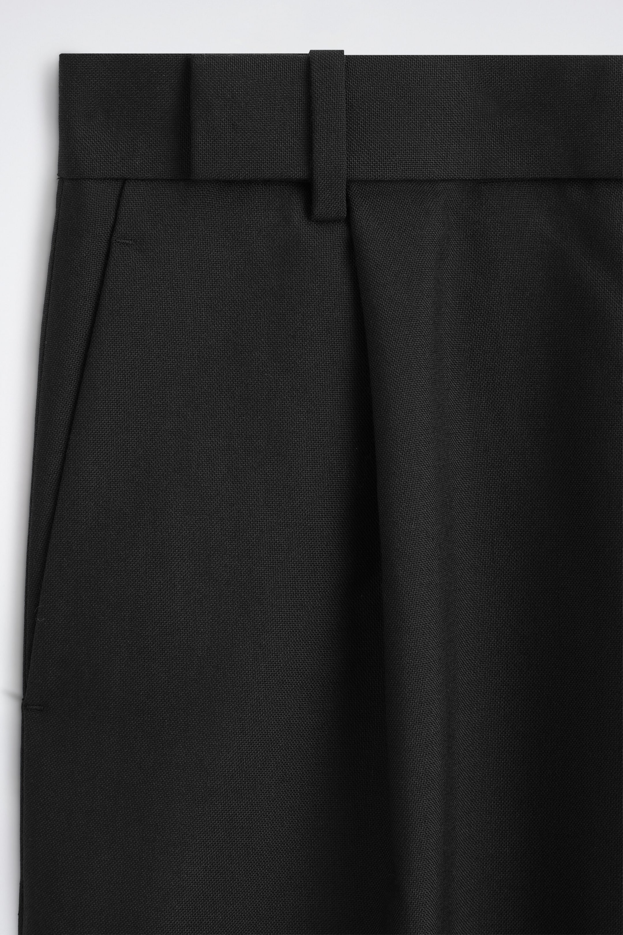 Eine Nahaufnahme von vorne der schwarzen Wollhose zeigt den strukturierten Bund mit einer breiten Gürtelschlaufe und einem integrierten Stoffgürtel. Eine einzelne Frontfalte sorgt für einen weichen Fall, während eine seitliche Eingrifftasche entlang der Naht des strukturierten Stoffes verläuft. Eine Nahaufnahme von vorne der schwarzen Wollhose zeigt den strukturierten Bund mit einer breiten Gürtelschlaufe und einem integrierten Stoffgürtel. Eine einzelne Frontfalte sorgt für einen weichen Fall, während eine seitliche Eingrifftasche entlang der Naht des strukturierten Stoffes verläuft.