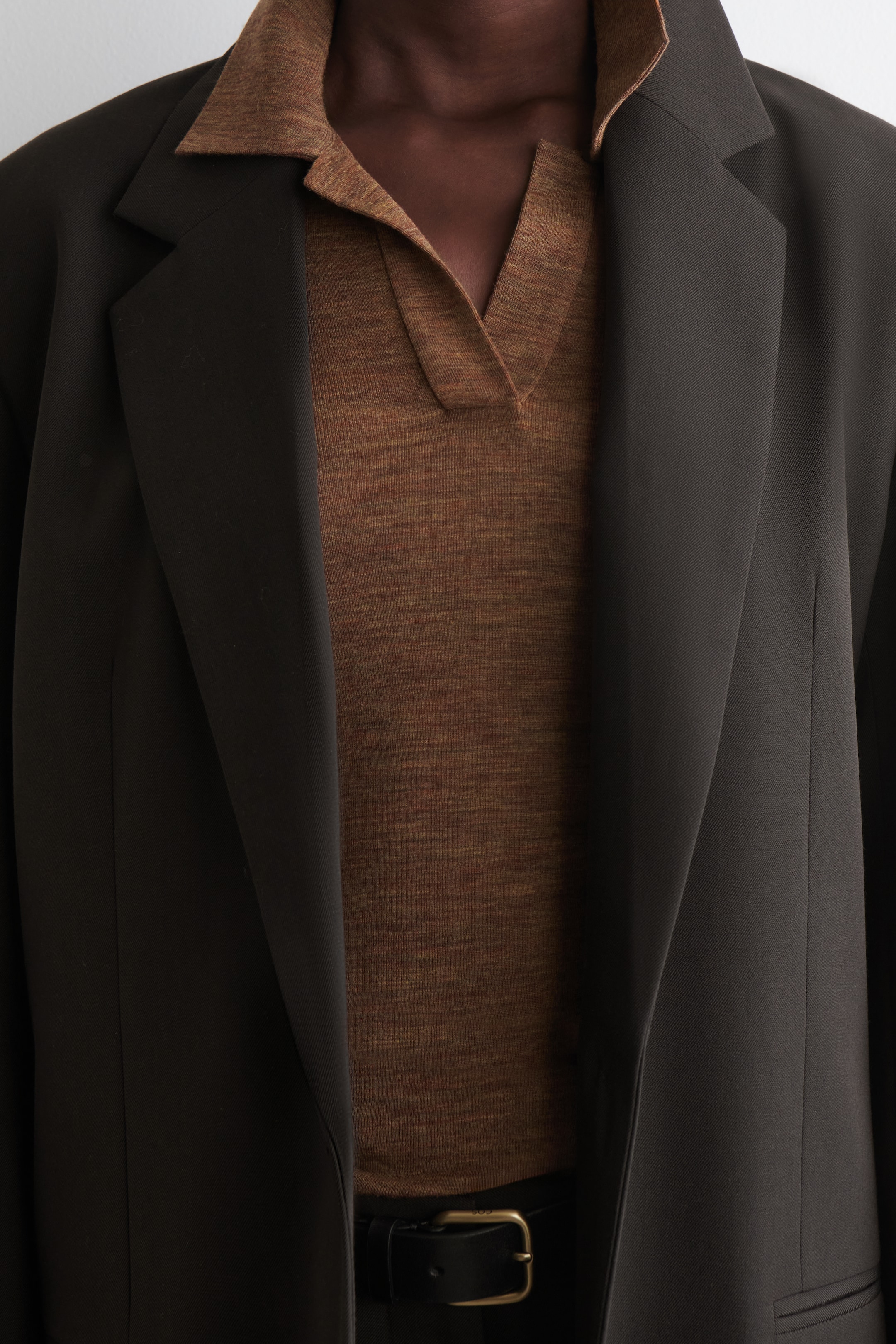 Visualizza immagine più grande: Un blazer scuro e aperto viene indossato, caratterizzato da ampi revers strutturati. Sotto, è visibile una polo in maglia marrone, con un colletto aperto e un'abbottonatura a V. Una cintura nera con una fibbia in ottone è anche visibile in vita.