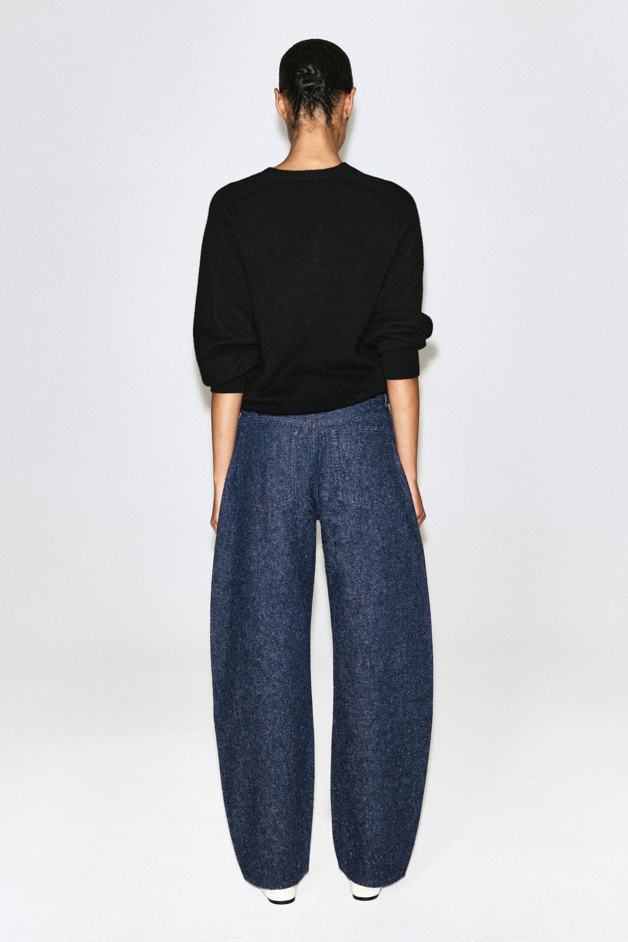 The dark blue, textured wide-leg denim pants are shown from the back, featuring a curved balloon silhouette and two back patch pockets. They are styled with a black, long-sleeve crewneck sweater. The dark blue, textured wide-leg denim pants are shown from the back, featuring a curved balloon silhouette and two back patch pockets. They are styled with a black, long-sleeve crewneck sweater.