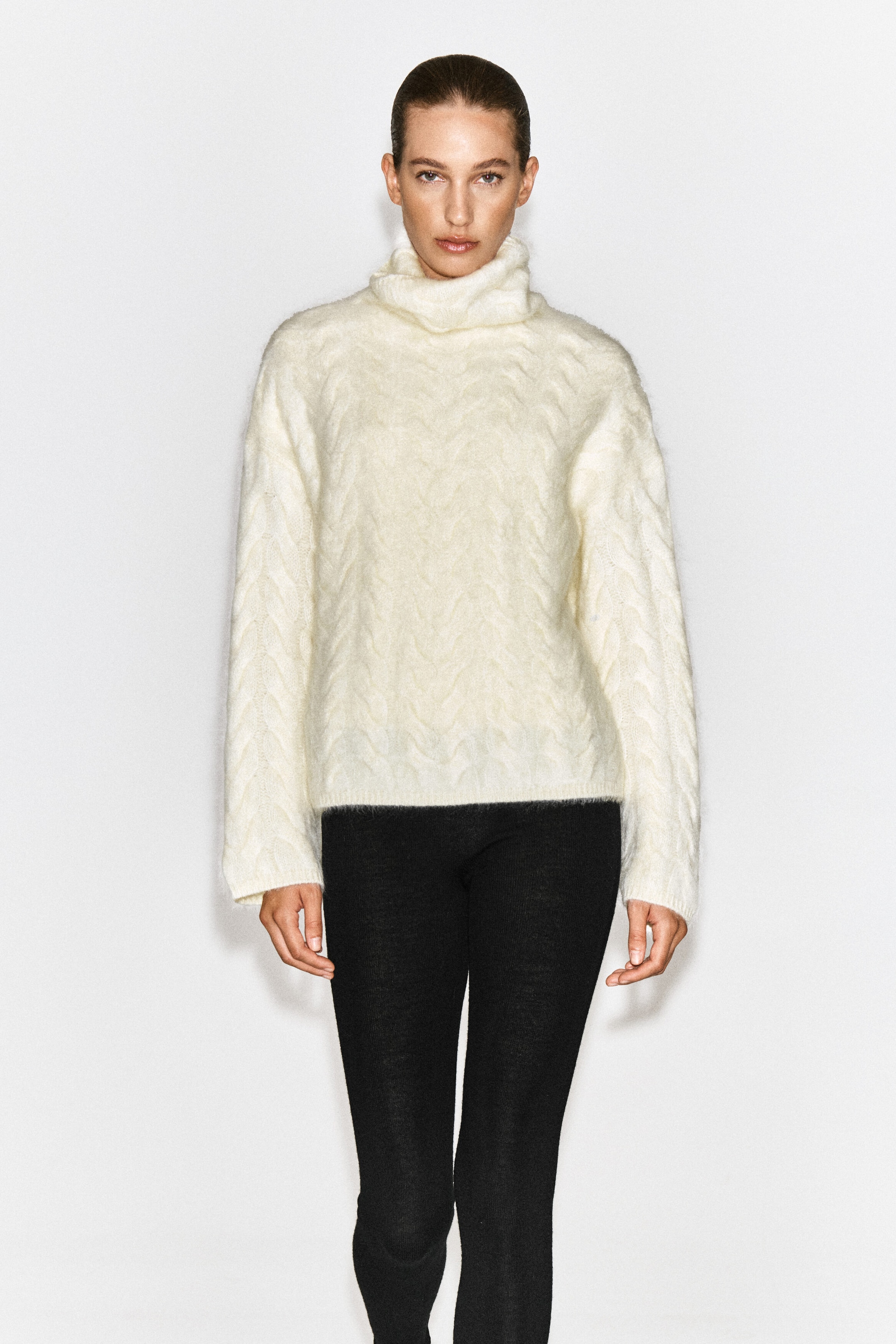 A cable-knit brushed cashmere funnel-neck jumper in an off-white shade is being worn by the model. The jumper features a textured cable pattern, a high funnel neck, and long sleeves, and is styled with black slim-fit trousers.