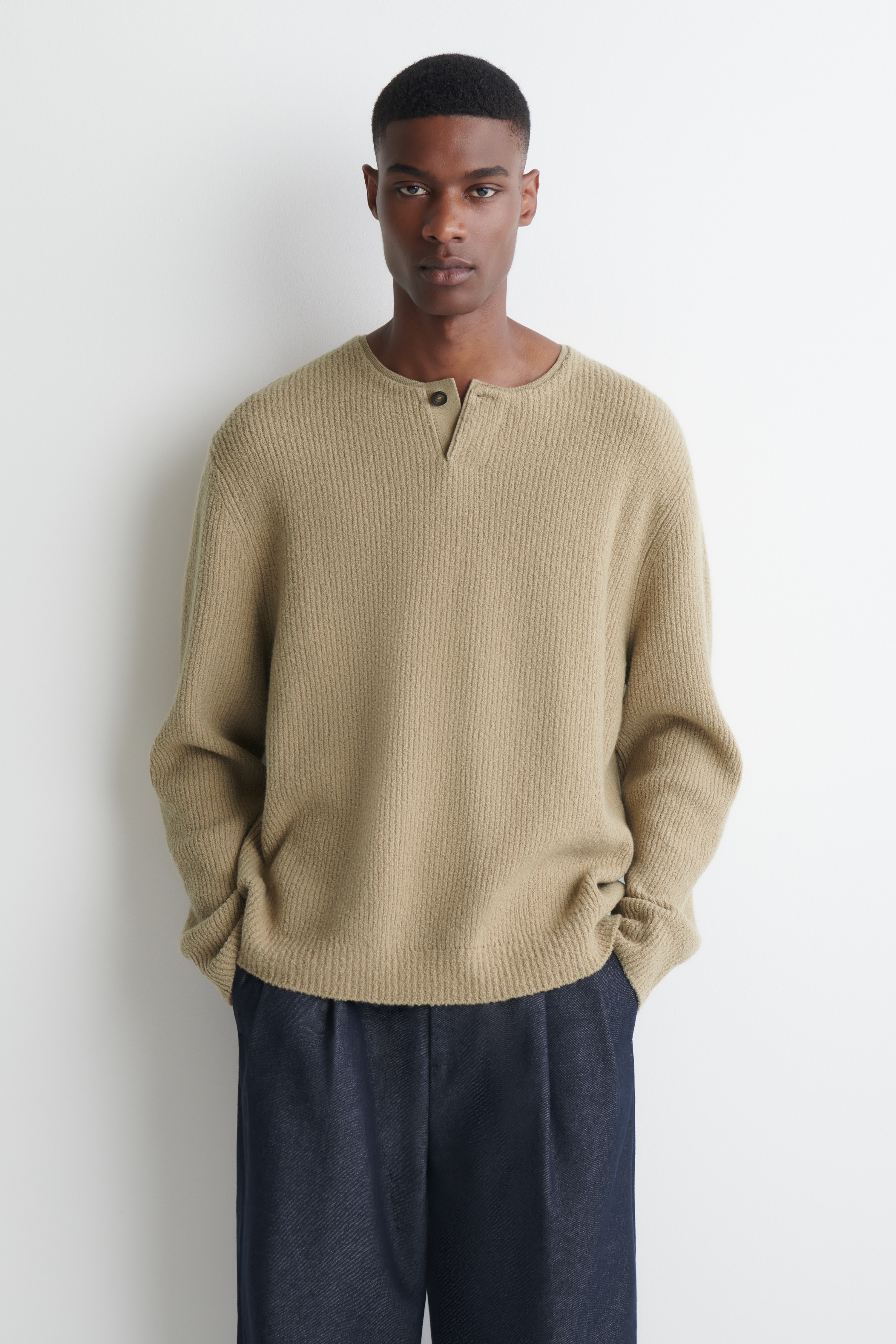 En beige ribbestrikket henley-genser er vendt forfra og viser en rund halsutringning med knappestolpe med Ã©n knapp og senkede skuldre. Genseren har lange ermer med ribbestrikkede mansjetter og en rett, ribbestrikket fald.