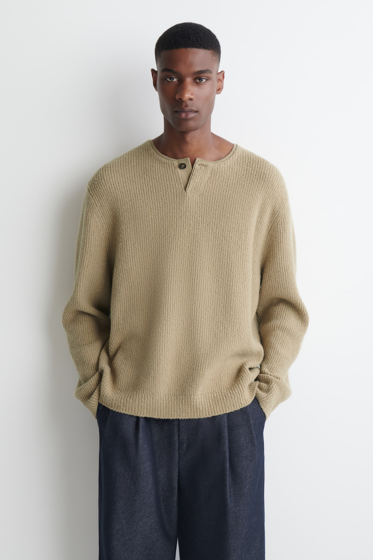 Un pull henley en maille cÃ´telÃ©e beige est portÃ© par le mannequin, avec une patte de boutonnage Ã  un seul bouton au col et une coupe dÃ©contractÃ©e. Ce pull Ã  manches longues est assorti Ã  un pantalon bleu marine foncÃ©. - 1
