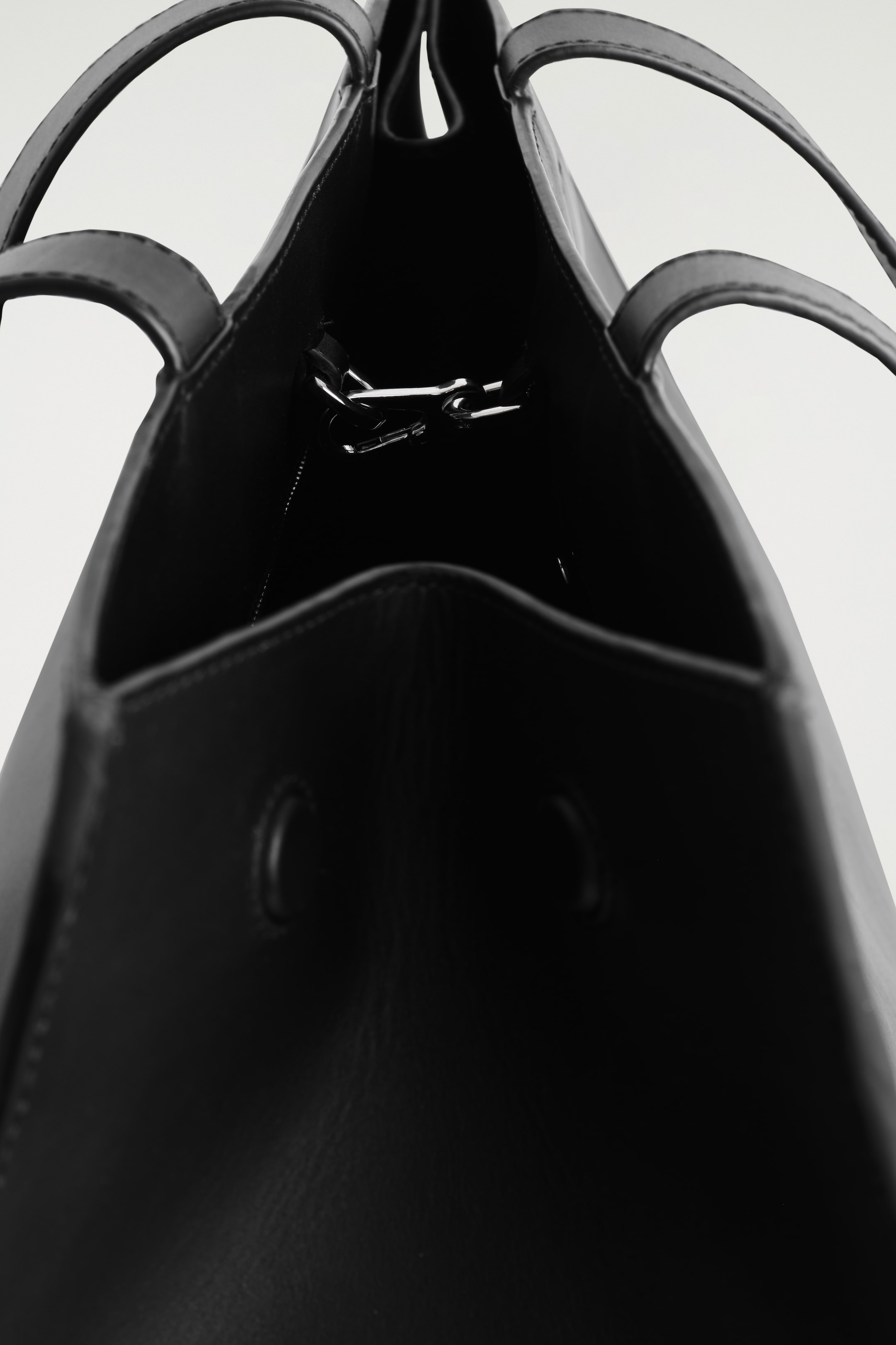 Visualizza immagine più grande: Una borsa tote in pelle nera viene vista dall'alto, mostrando il suo interno aperto con due manici superiori in pelle nera che si arcuano verso l'alto. Una chiusura a clip in metallo color argento si estende attraverso l'apertura della borsa, e una tasca interna con un'apertura arrotondata è visibile lungo la parete interna anteriore.