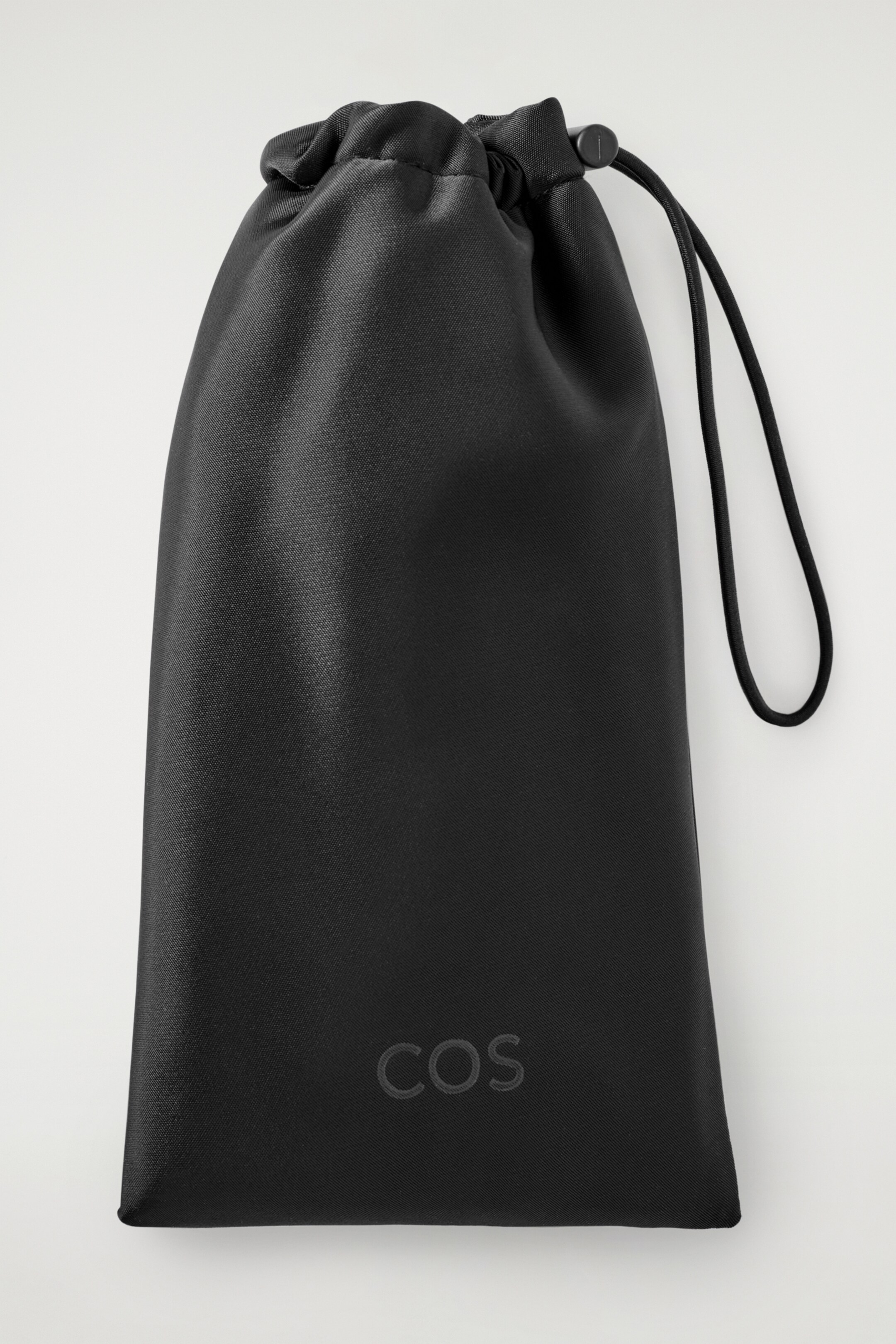 View larger image: A black drawstring pouch is presented front-on, featuring a gathered top closure with a black cord lock. The finely woven fabric pouch is displaying a subtle, tone-on-tone "COS" logo at its lower front.