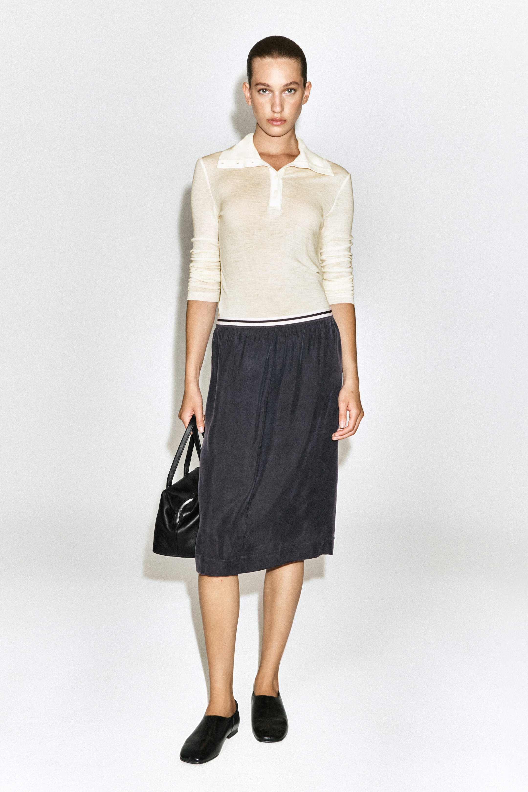 A light beige long-sleeve polo shirt, a dark gray knee-length skirt with a striped elastic waistband, and black flat slip-on shoes are being worn by the model, who is also holding a black structured handbag. A light beige long-sleeve polo shirt, a dark gray knee-length skirt with a striped elastic waistband, and black flat slip-on shoes are being worn by the model, who is also holding a black structured handbag.