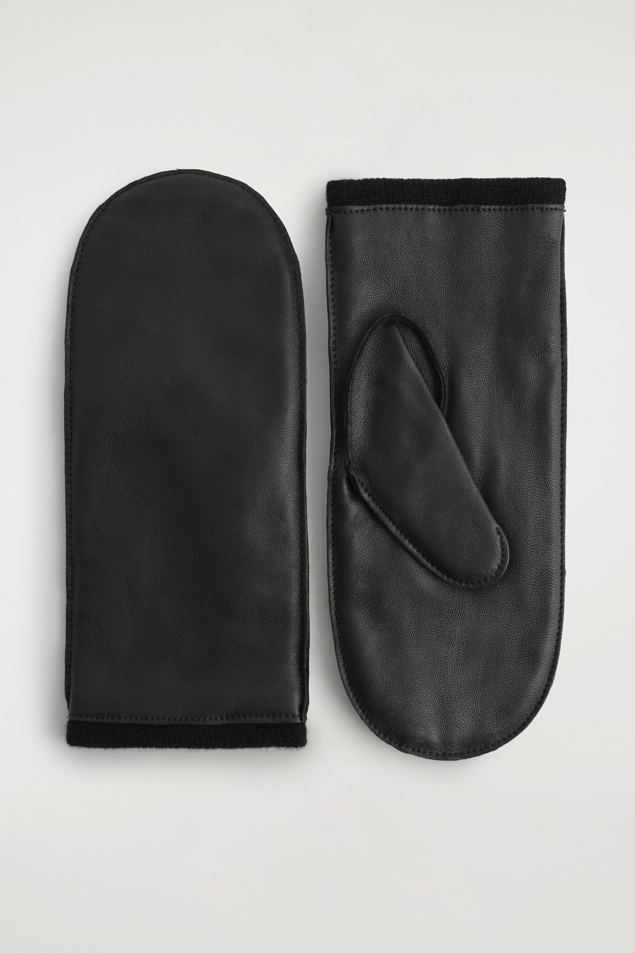A pair of black leather mittens is laid flat, with one mitten displaying its smooth back and the other showing its palm side with an integrated thumb piece. Both mittens feature a finely textured finish and a black ribbed knit cuff.