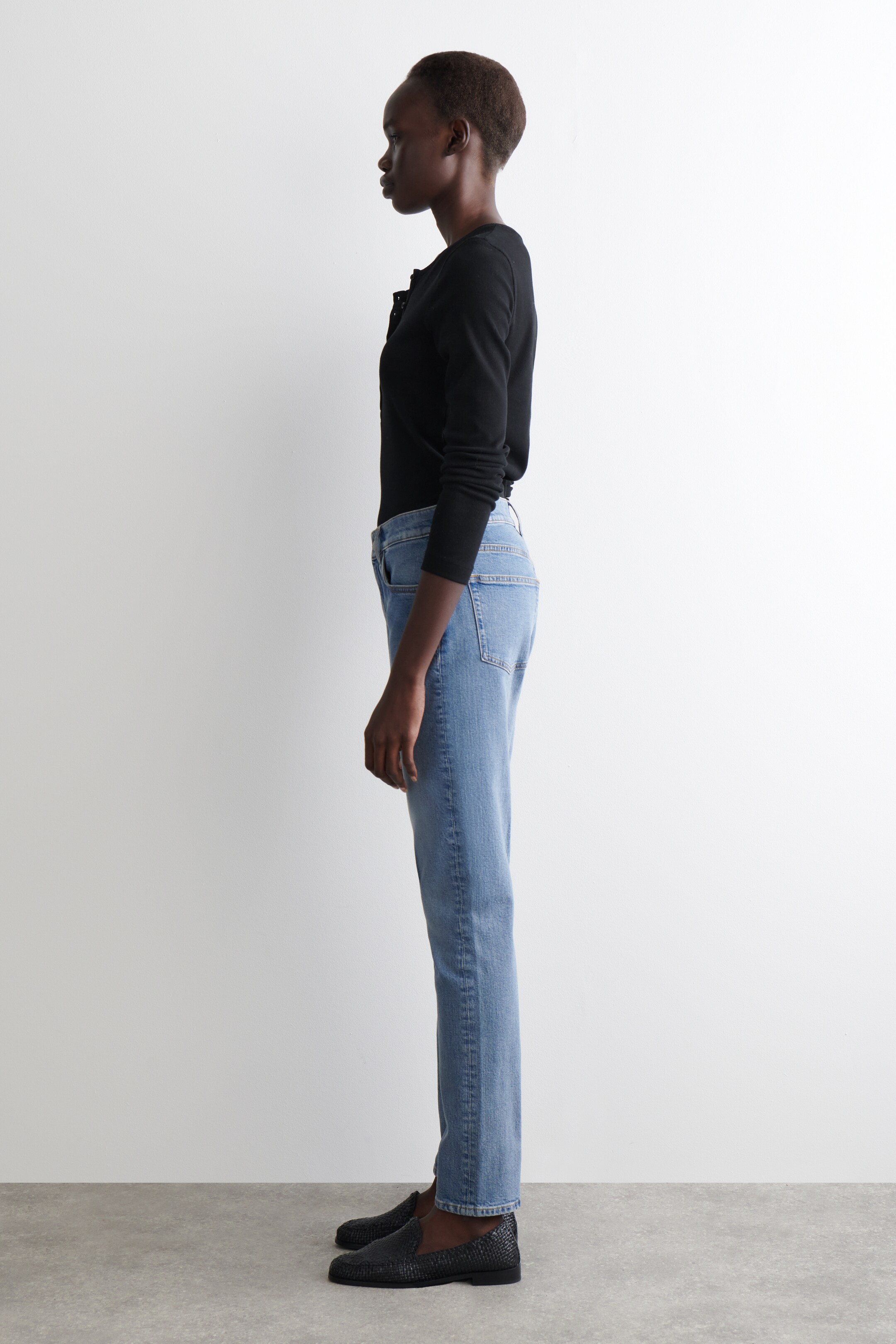 Agrandir l'image: Une tenue complète est présentée de profil, avec un haut noir à manches longues qui est ajusté et rentré dans un jean bleu clair à taille haute. Le jean présente une coupe droite, et des mocassins noirs texturés complètent l'ensemble.
