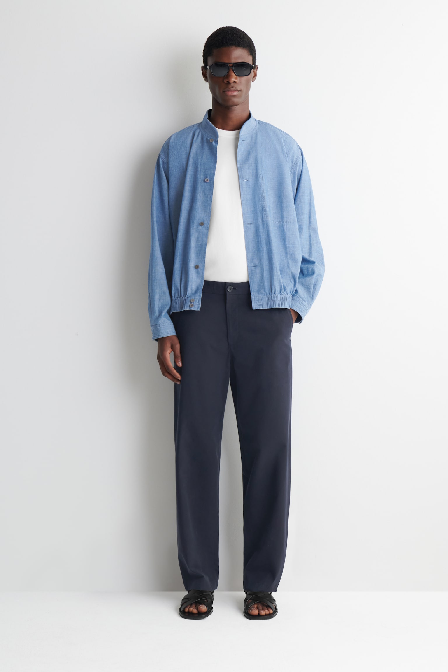 The model is wearing a blue chambray bomber jacket. It boasts a band collar, button-front fastening, elasticated cuffs, and an elasticated hem, and is layered over a white t-shirt. - 1