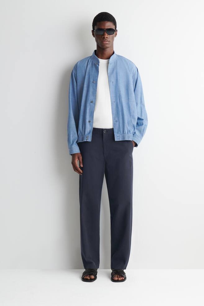 Een blauw chambray bomberjack wordt gedragen door het model. Het heeft een bandkraag, een knoopsluiting aan de voorkant, elastische manchetten en een elastische zoom, en is gelaagd over een wit t-shirt. - 1