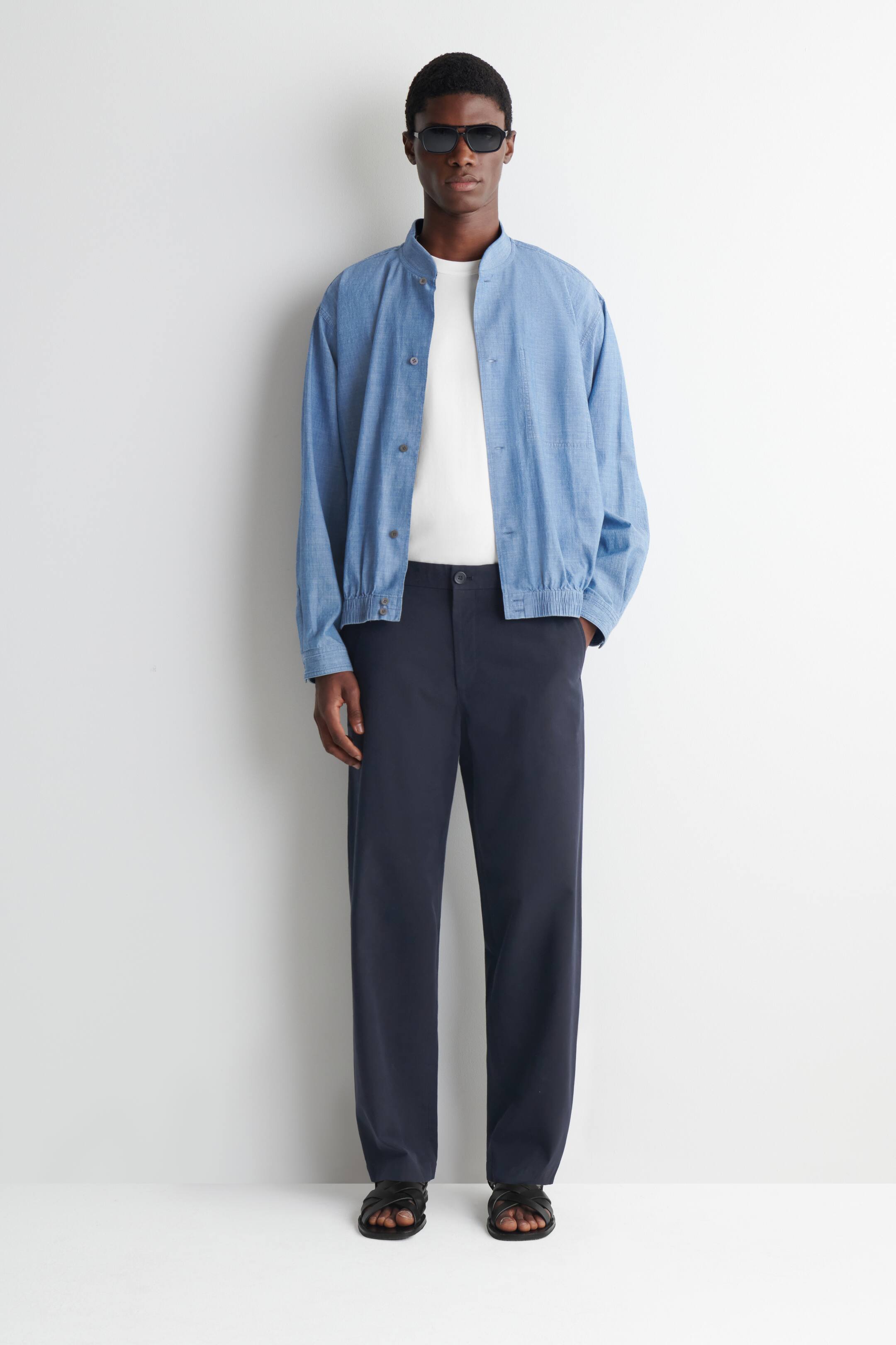 Agrandir l'image: Le mannequin porte un blouson bomber en chambray bleu. Il se distingue par un col à bande, une fermeture à boutons sur le devant, des poignets élastiqués et un ourlet élastiqué, et est superposé sur un t-shirt blanc.