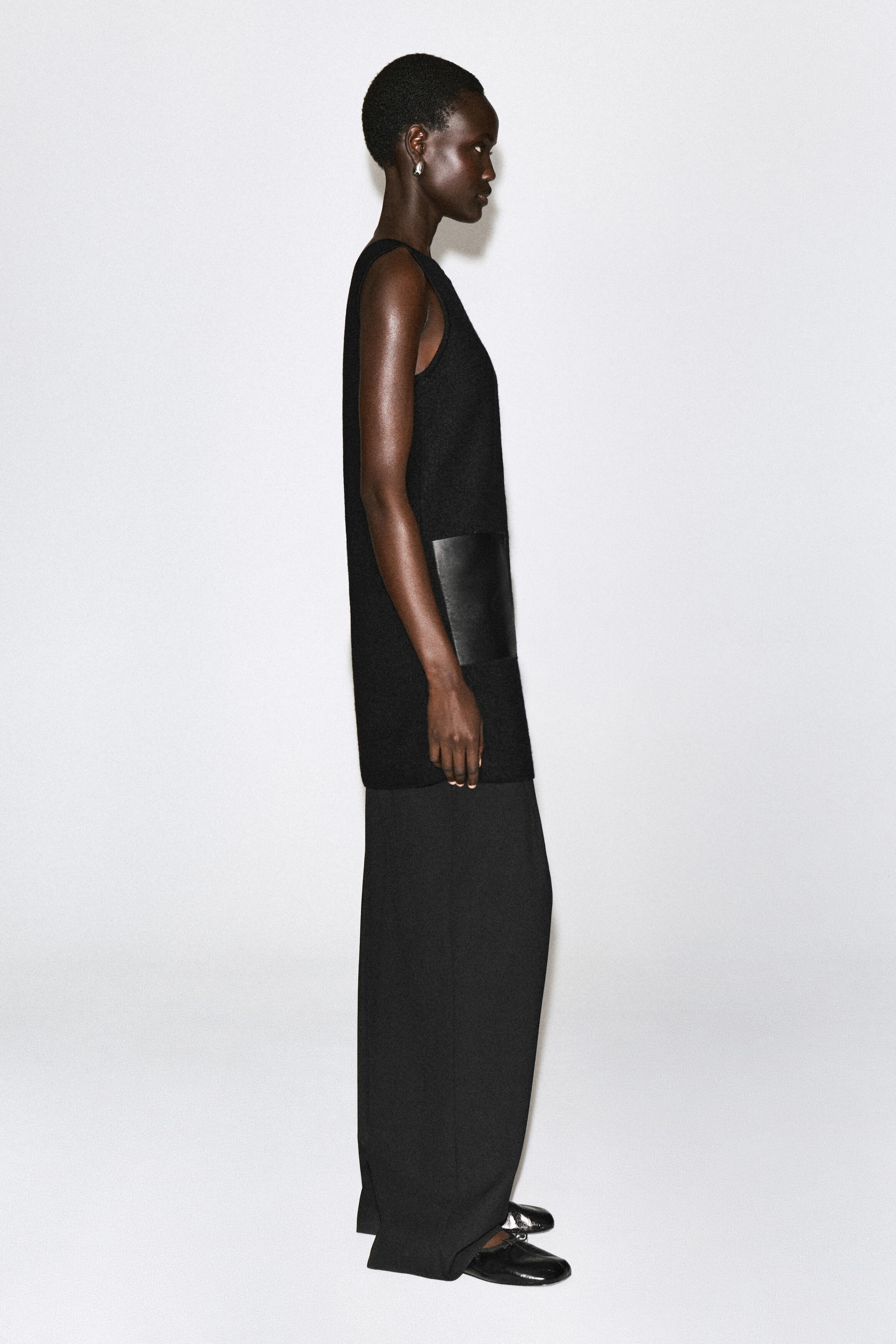 A sleeveless black boiled wool mini dress is shown in right-side profile, featuring a smooth black leather patch pocket on the side and layered over wide-leg black trousers to highlight its structured, straight silhouette. A sleeveless black boiled wool mini dress is shown in right-side profile, featuring a smooth black leather patch pocket on the side and layered over wide-leg black trousers to highlight its structured, straight silhouette.