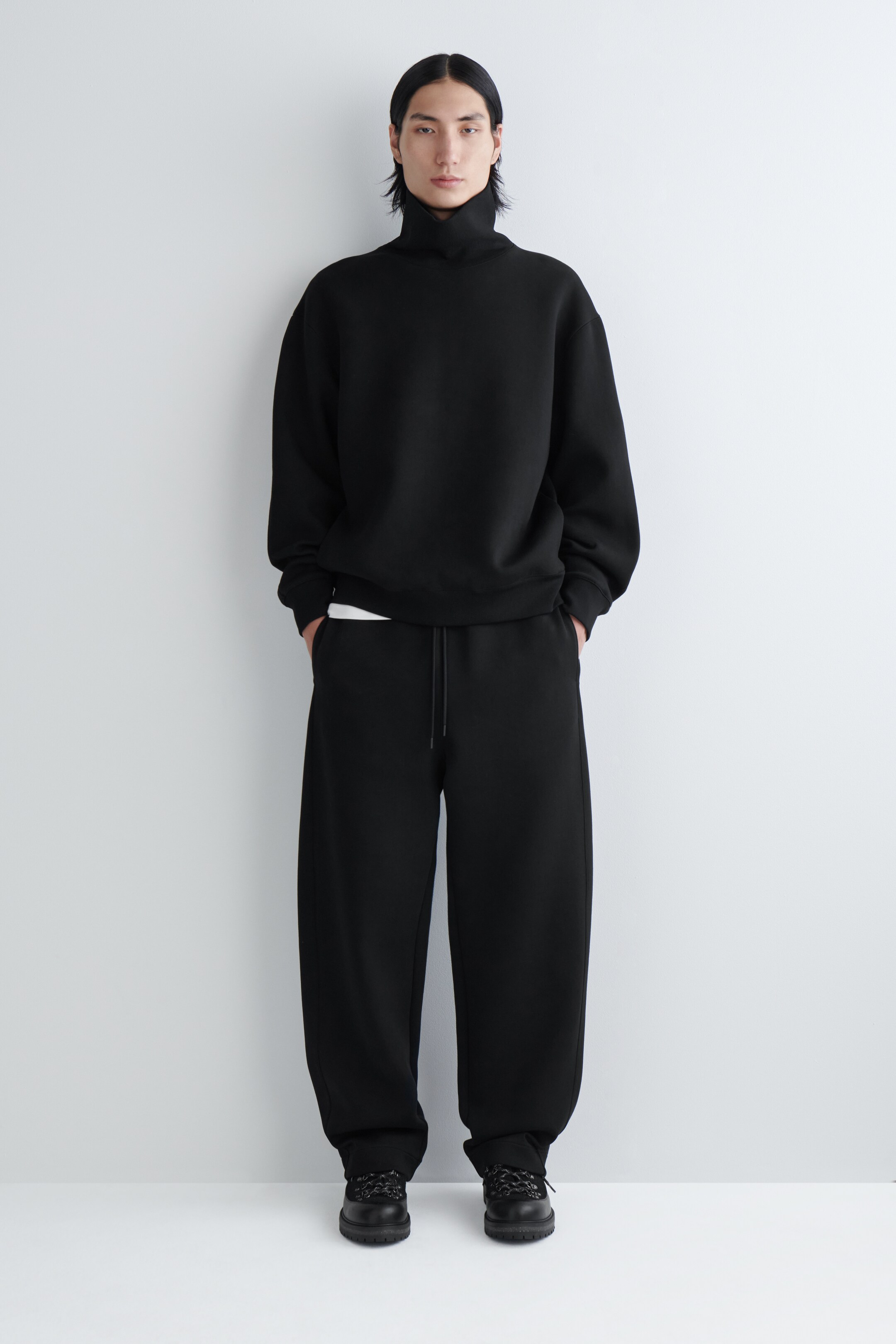 Agrandir l'image: Un sweat-shirt noir à col roulé ample et un pantalon de survêtement noir à jambes larges sont présentés de face. Le sweat-shirt surdimensionné présente une couche blanche dépassant de son ourlet, et le pantalon de survêtement est doté d'une taille à cordon de serrage et s'accumule sur des chaussures noires épaisses.