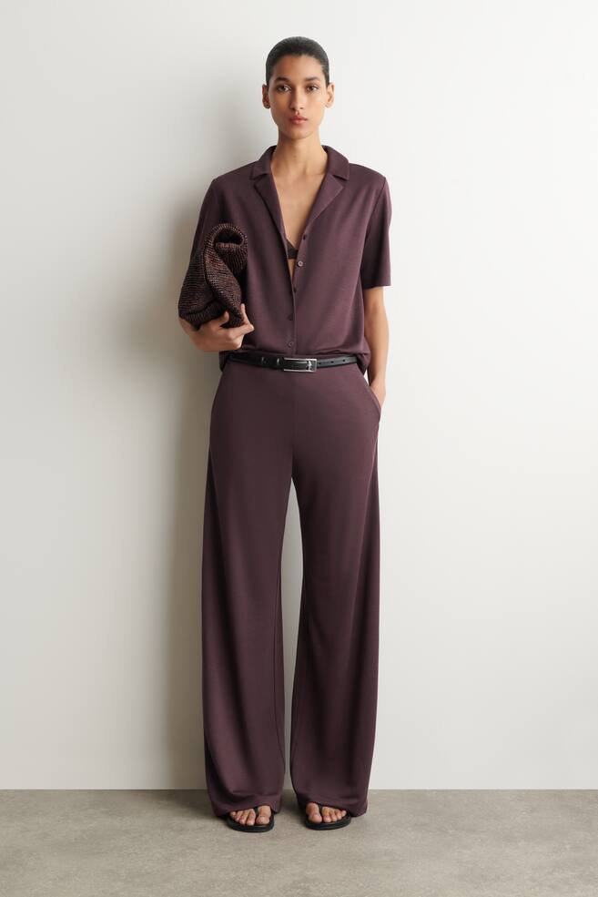 A full-front view showcases a deep plum short-sleeved shirt paired with matching wide-leg trousers. The shirt features a camp collar and front button placket, revealing a black bralette, while the wide-leg trousers are cinched at the waist with a black belt. - 1