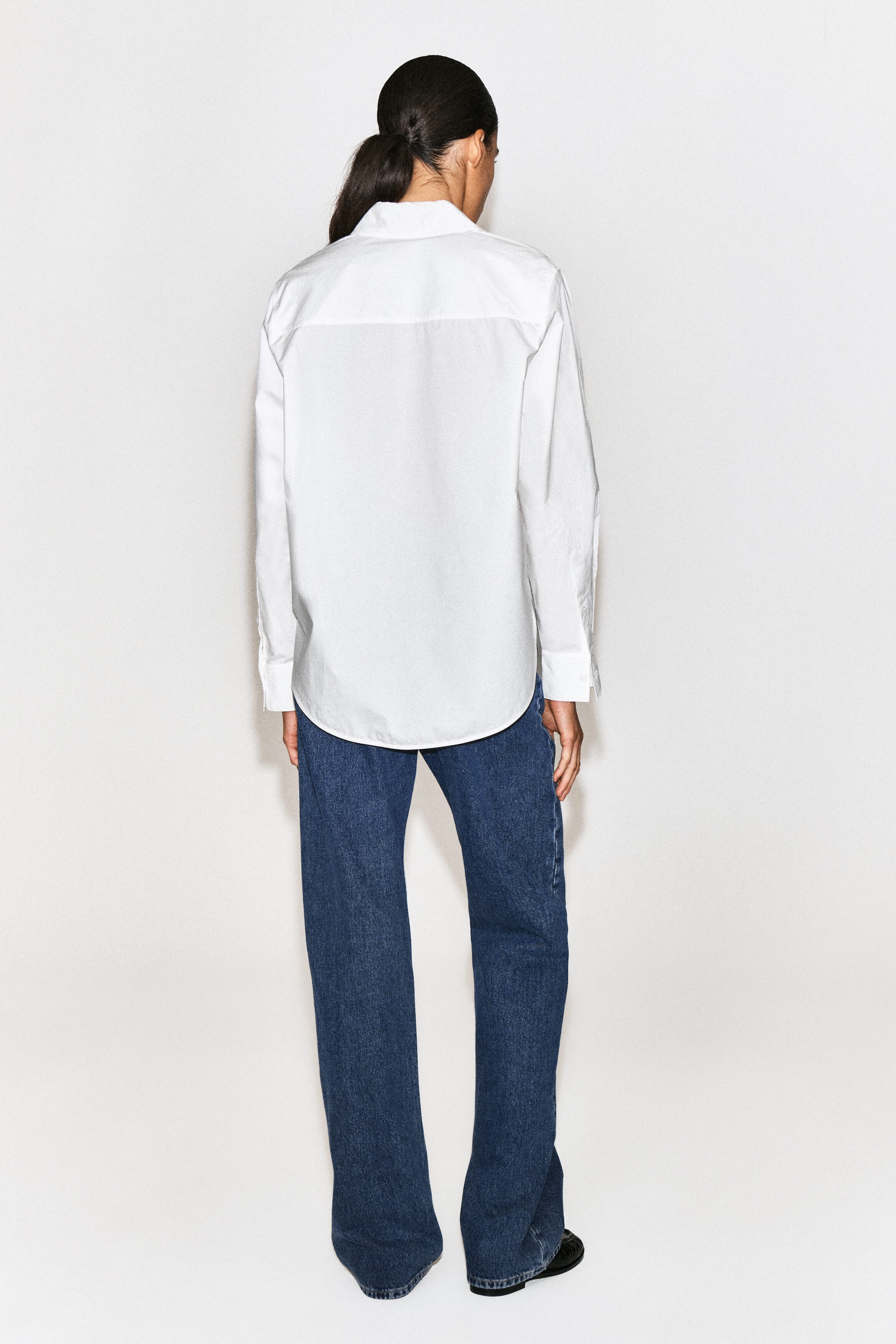 The classic cotton poplin shirt is shown from the back, highlighting its relaxed fit, straight back yoke, long sleeves with buttoned cuffs, and a curved hem that drapes over blue jeans. The classic cotton poplin shirt is shown from the back, highlighting its relaxed fit, straight back yoke, long sleeves with buttoned cuffs, and a curved hem that drapes over blue jeans.