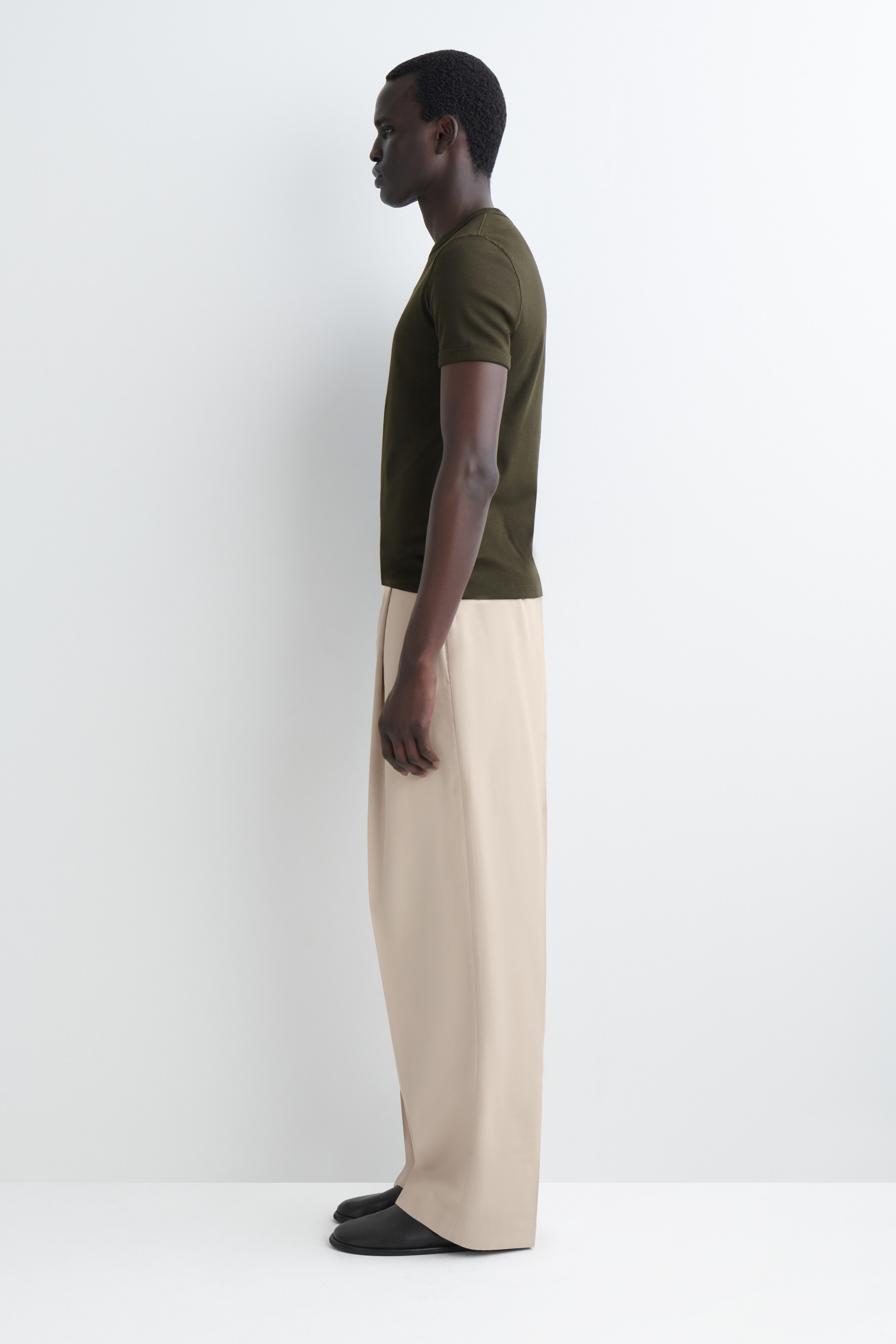 Visualizza immagine più grande: Una maglietta a maniche corte verde oliva scuro è abbinata a pantaloni beige chiaro a gamba larga, presentata di profilo. I pantaloni cadono sopra scarpe basse nere.