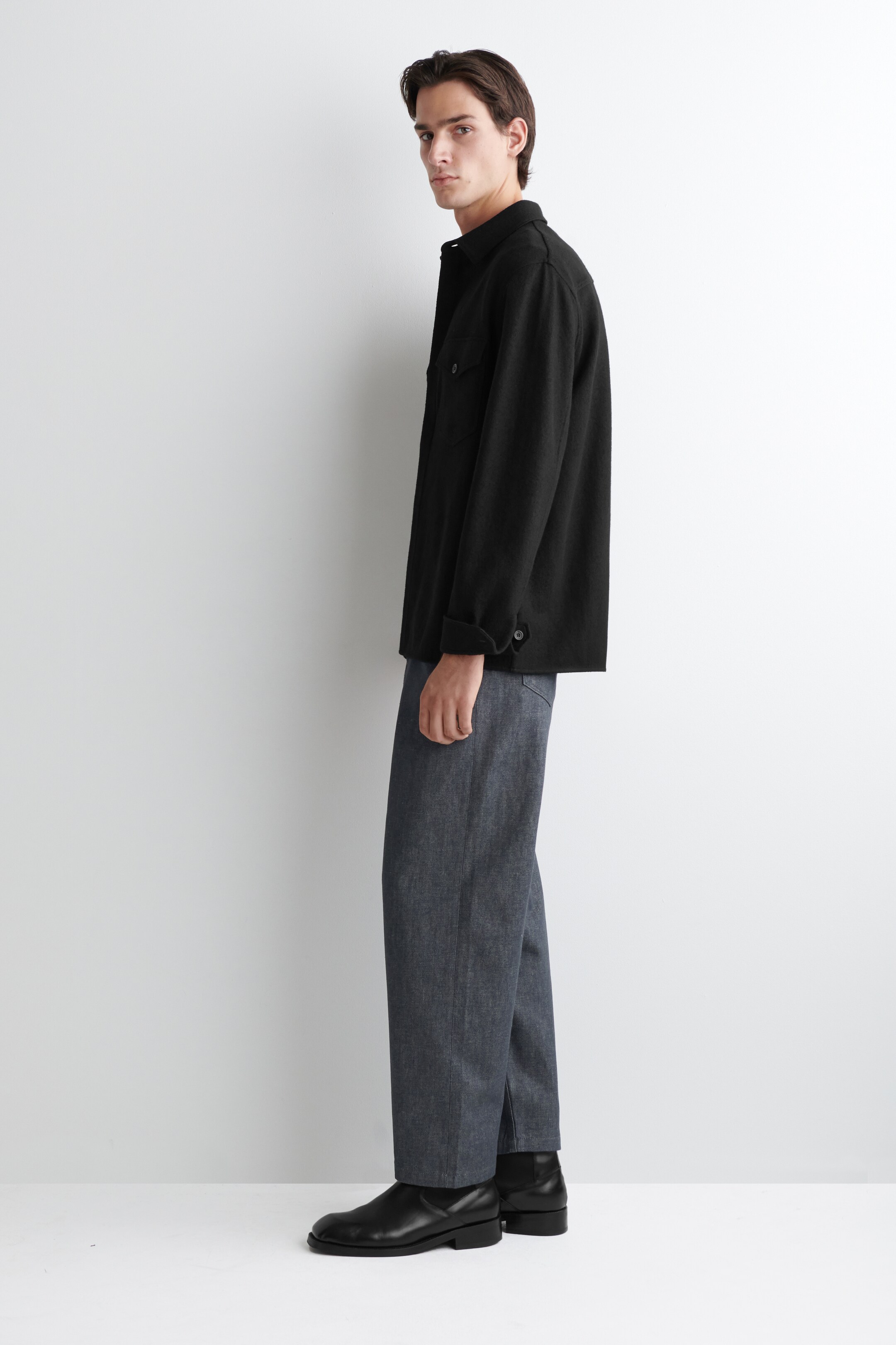 Visualizza immagine più grande: Una overshirt nera viene presentata in un profilo laterale, caratterizzata da un colletto a punta, una tasca sul petto visibile e polsini arrotolati con bottoni. Viene indossata con pantaloni grigio scuro a gamba larga che coprono stivaletti neri.