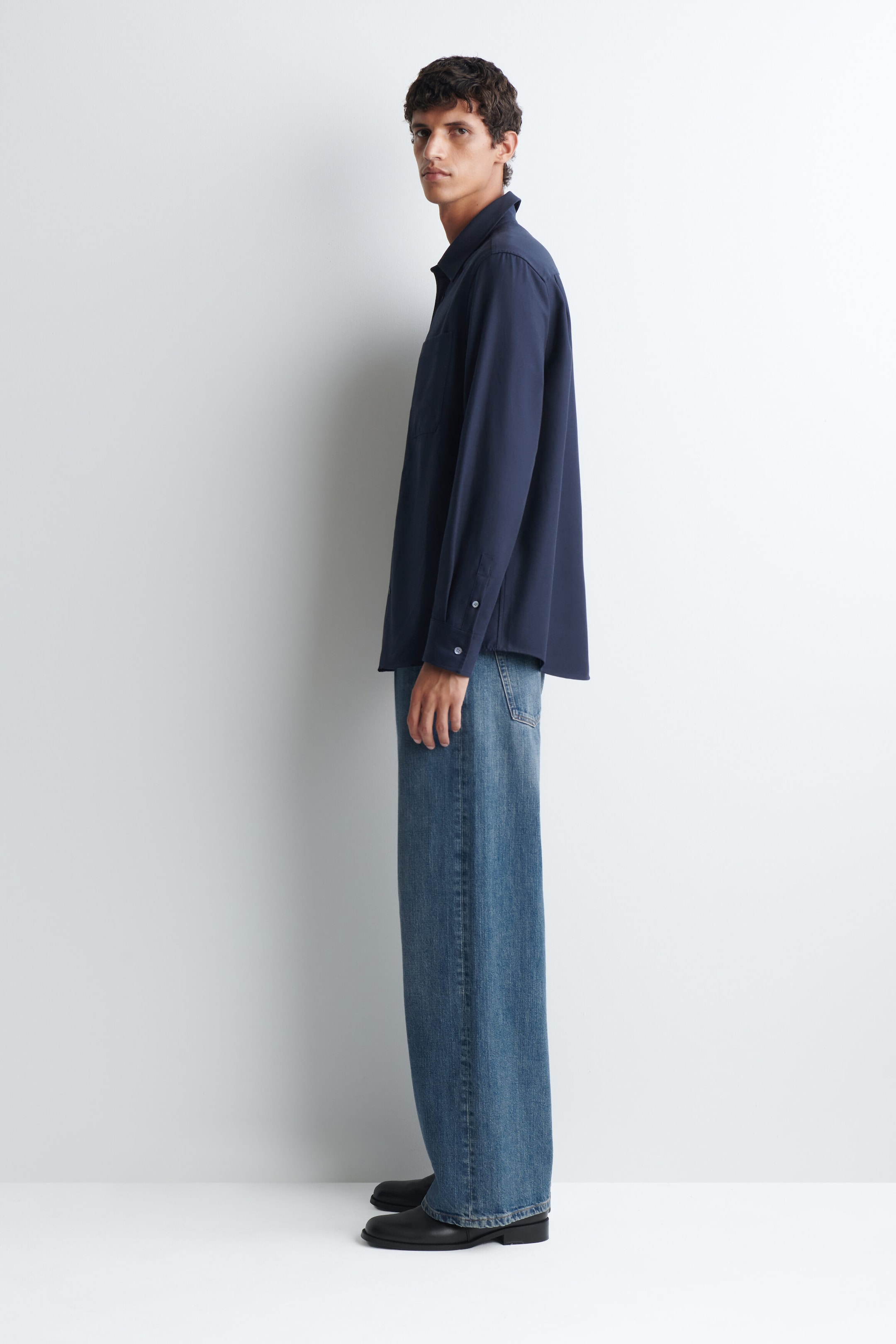 The relaxed twill shirt in navy blue is being worn by the model, who is standing in profile. This long-sleeved shirt features a classic collar and buttoned cuffs, and is styled with wide-leg blue jeans.