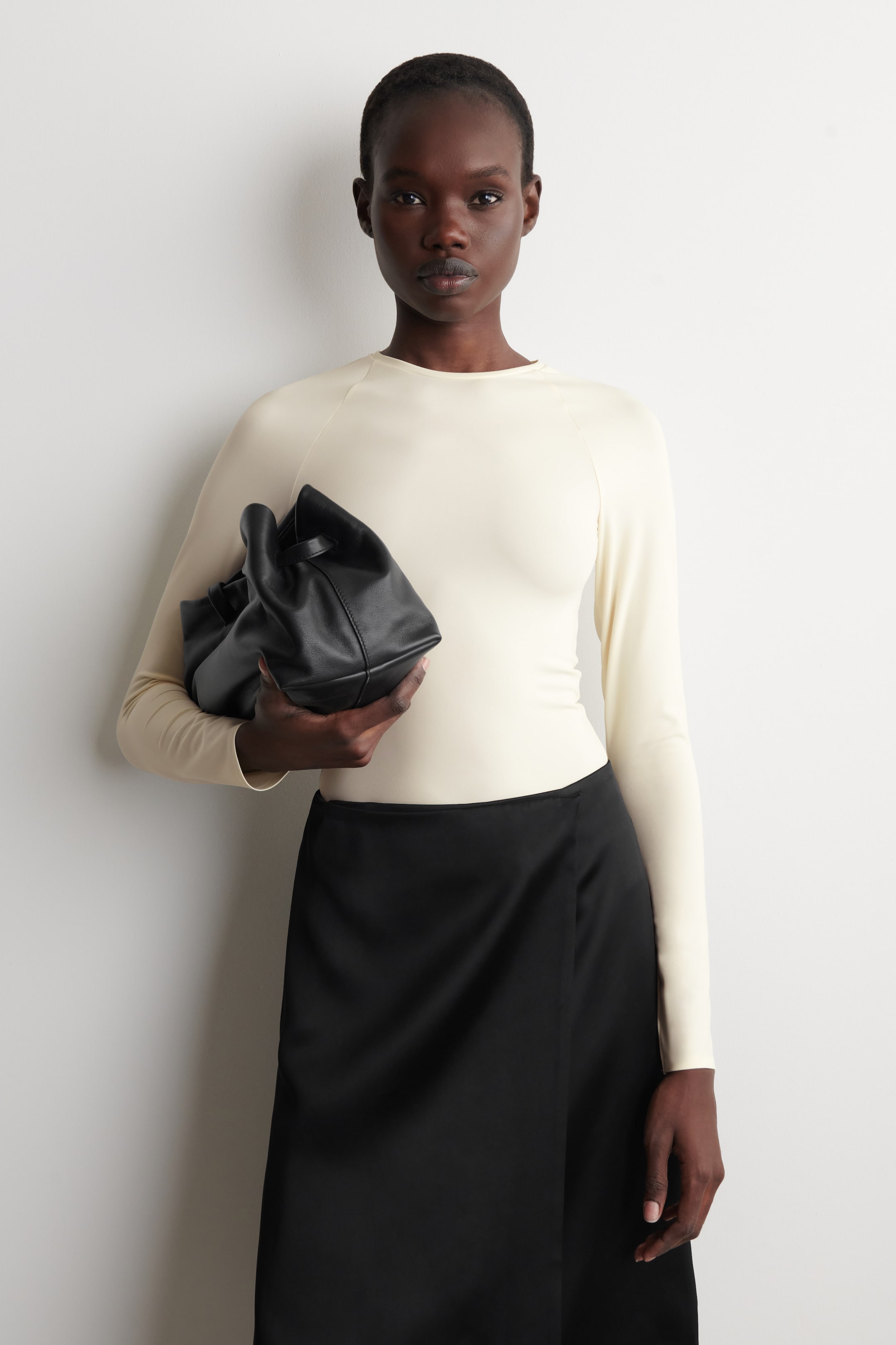 Vezi imaginea mai mare: Un top de culoare crem, cu croială slim-fit, este prezentat din față, având un decolteu rotund și mâneci lungi raglan. Topul apare neted și mulat, fiind asortat cu o fustă neagră cu înfășurare.