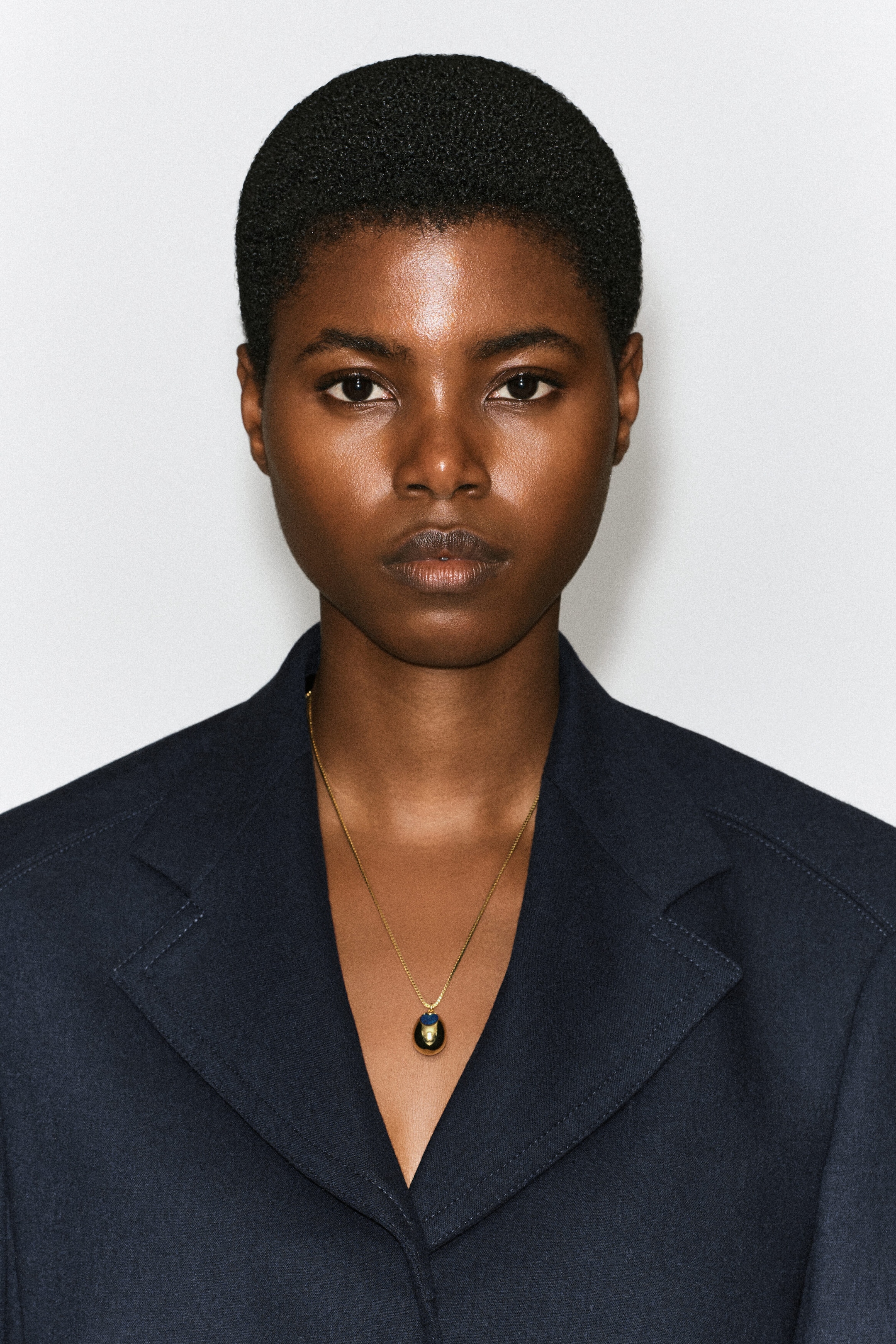 A lapis lazuli pendant necklace, featuring a delicate gold chain and a rounded gold pendant set with a blue stone, is worn by the model. The necklace rests against the collarbone and is styled with a dark blue blazer. A lapis lazuli pendant necklace, featuring a delicate gold chain and a rounded gold pendant set with a blue stone, is worn by the model. The necklace rests against the collarbone and is styled with a dark blue blazer.