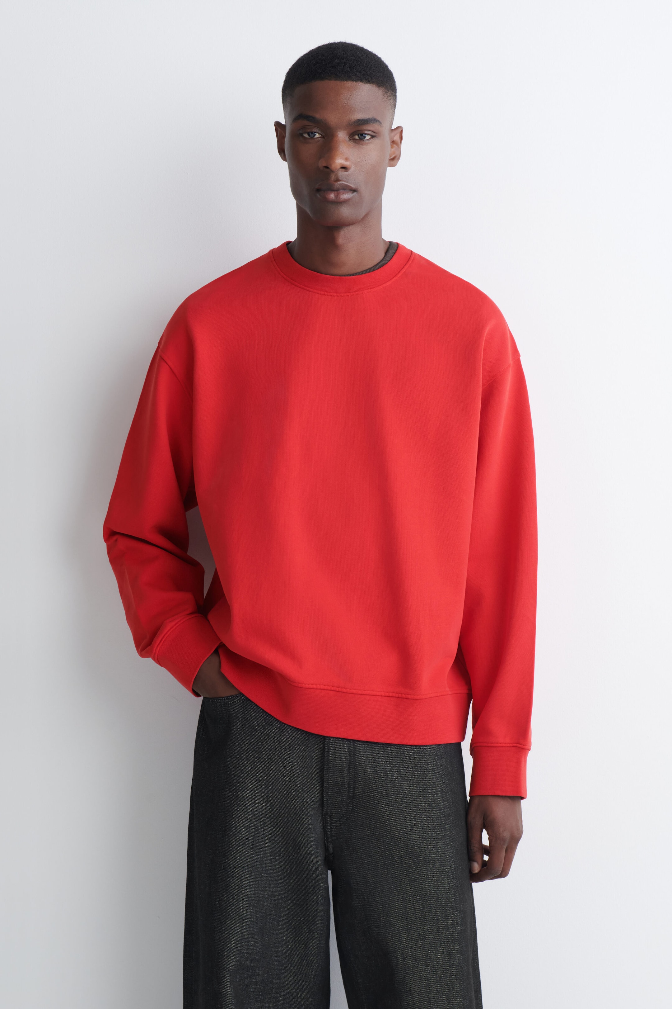 A bright red crew-neck sweatshirt with ribbed trims is being worn by the model. The relaxed-fit sweatshirt is paired with dark grey trousers, with the model placing one hand in a pocket. A bright red crew-neck sweatshirt with ribbed trims is being worn by the model. The relaxed-fit sweatshirt is paired with dark grey trousers, with the model placing one hand in a pocket.