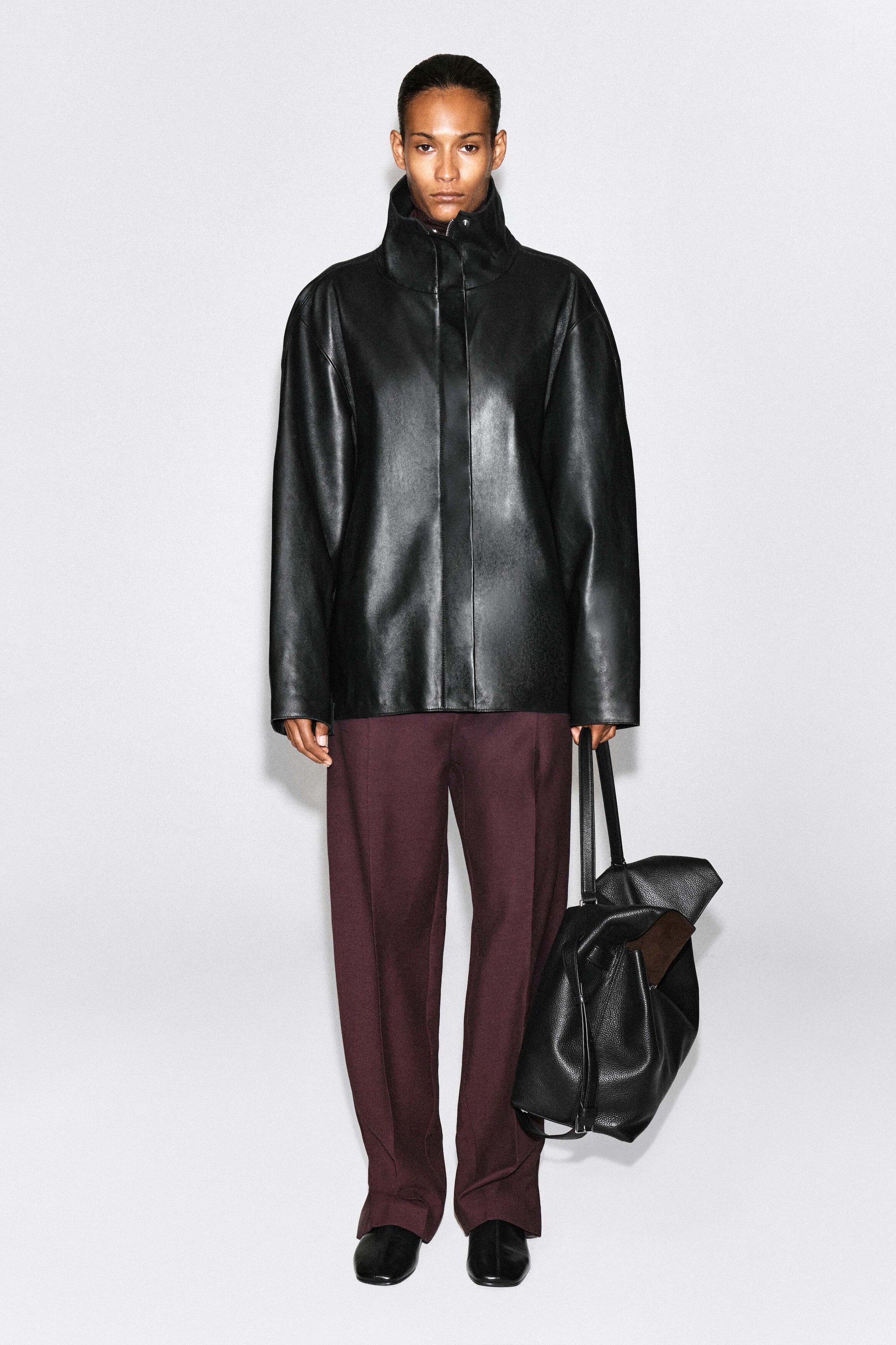 Pleated-panel straight-leg pants in a deep burgundy color are worn by the model, featuring a sharp center crease along each leg. The pants are styled with a high-collar black leather jacket and black shoes. Pleated-panel straight-leg pants in a deep burgundy color are worn by the model, featuring a sharp center crease along each leg. The pants are styled with a high-collar black leather jacket and black shoes.