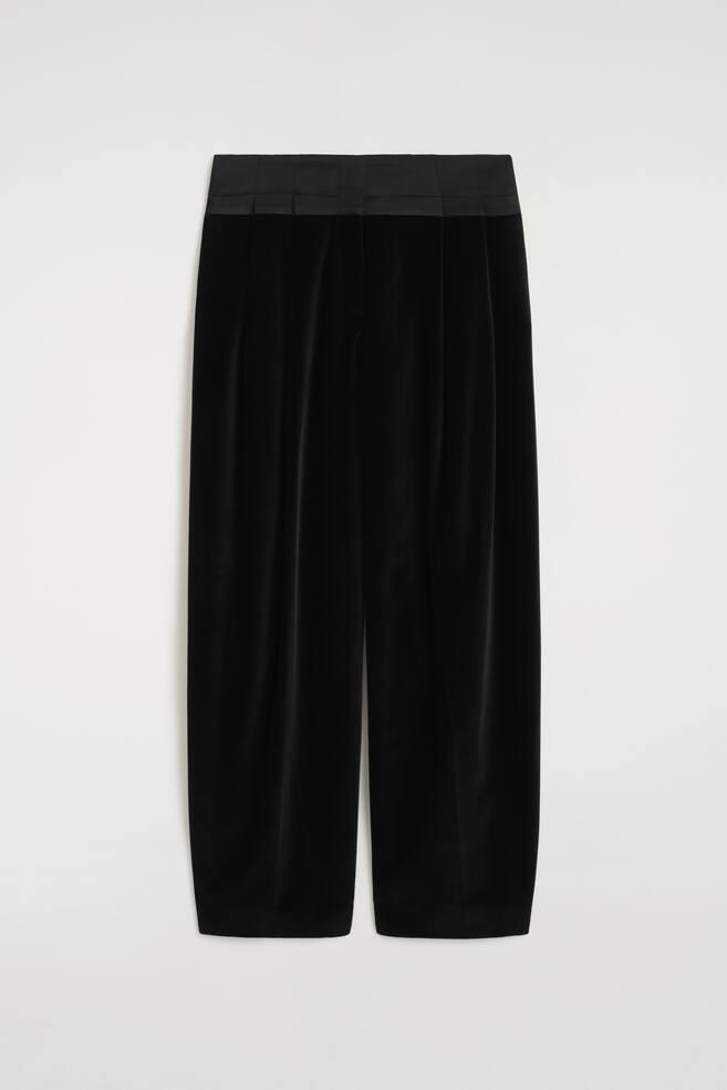 A pair of black satin-trimmed velvet barrel-leg trousers is shown from the front, featuring a wide, structured satin waistband with subtle pleats that create volume. The trousers showcase a relaxed, curved barrel-leg silhouette crafted from plush black velvet, tapering slightly at the hem. - 1