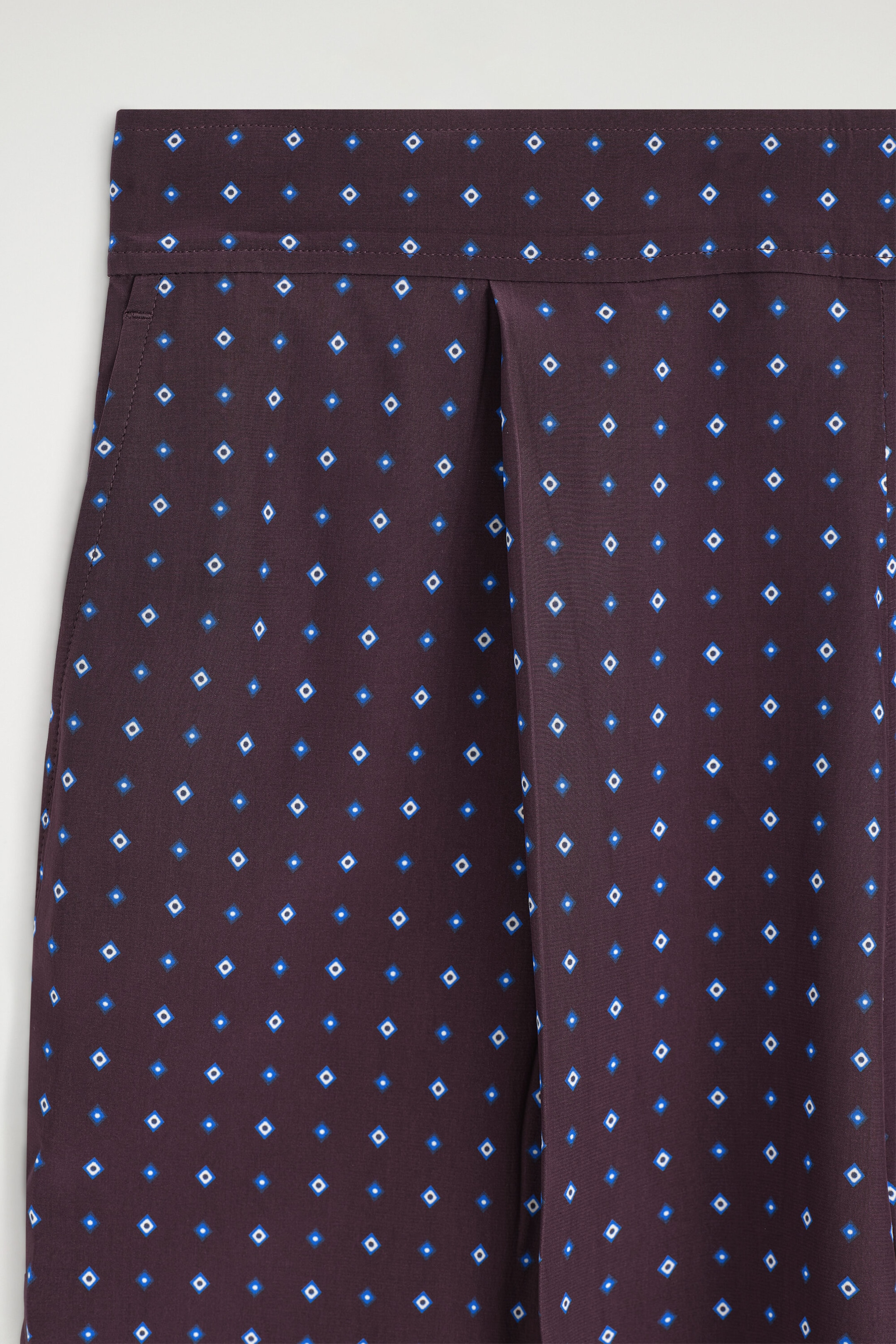 Ce pantalon droit à motif losanges présente un tissu bordeaux profond orné de petits losanges géométriques bleus et blancs répétés, en gros plan de face. Le modèle comporte une taille plate, une poche latérale passepoilée et un pli structuré sur le devant.