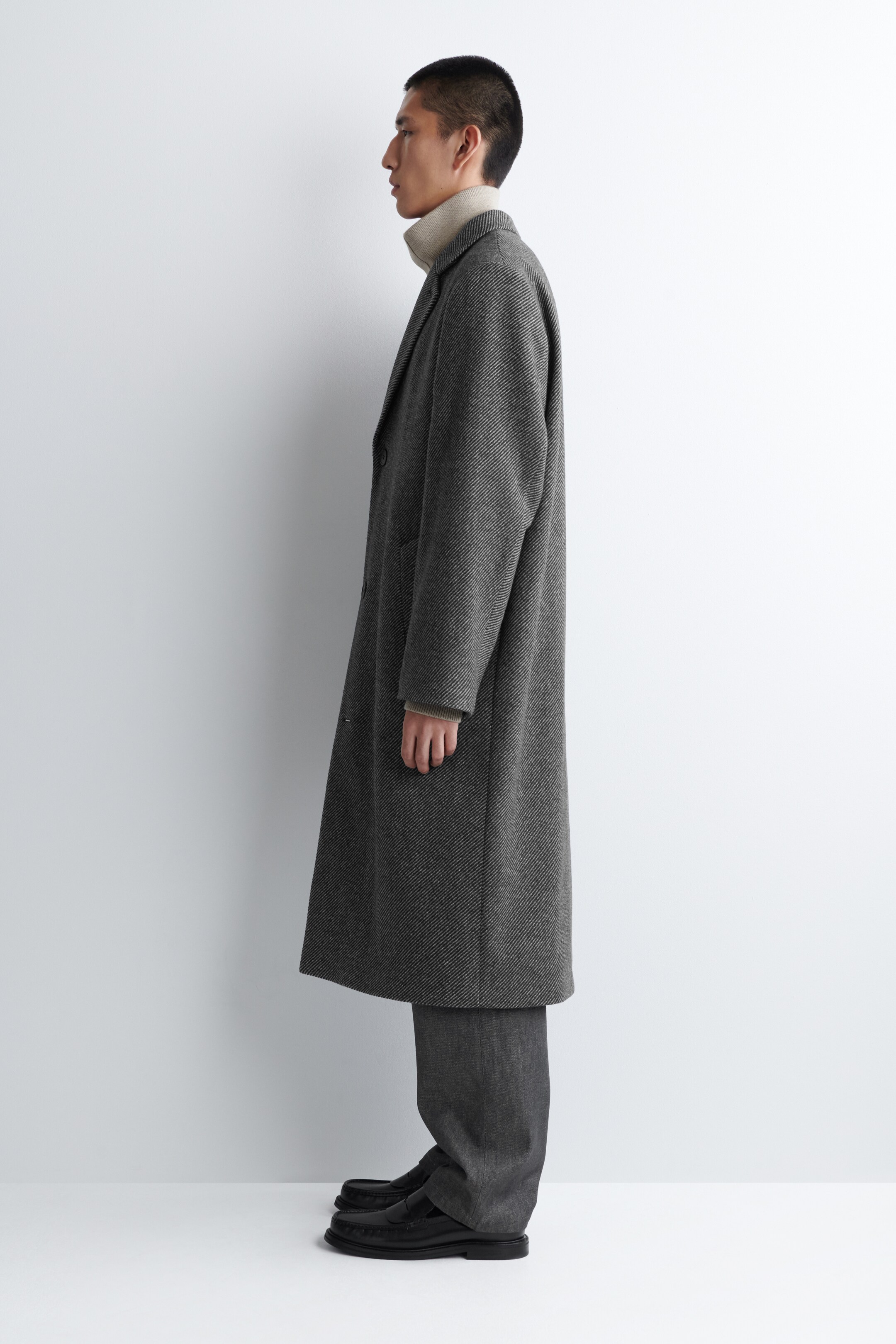 Agrandir l'image: Un long manteau gris foncé est présenté de côté, avec une texture de tissage en diagonale, des revers crantés et deux boutons visibles à l'avant. Un pull à col roulé de couleur claire est en dessous, visible au niveau du cou et des manches, tandis que des pantalons sombres apparaissent sous l'ourlet du manteau.