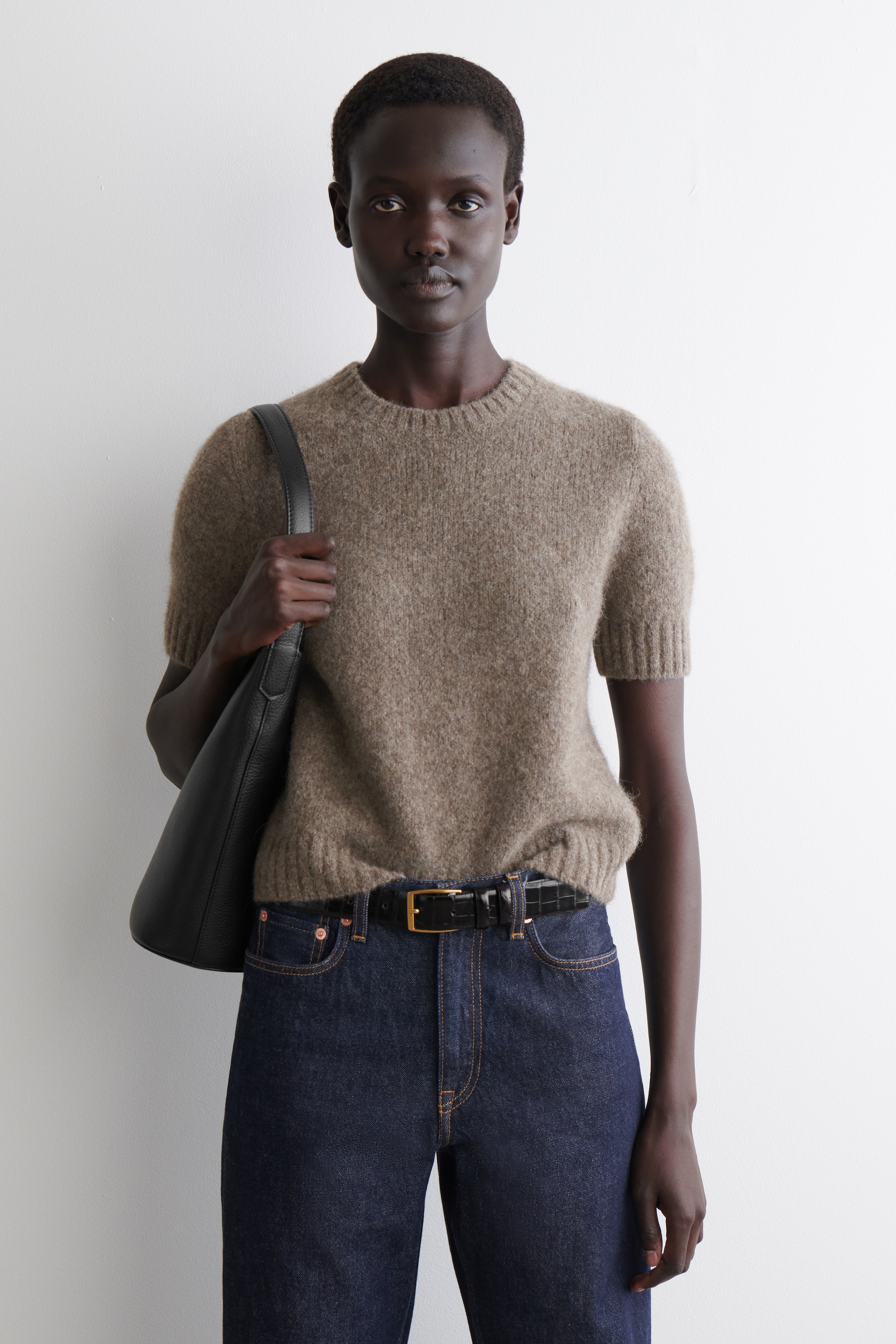 Agrandir l'image: Un pull à manches courtes en tricot duveteux de couleur brun clair est présenté en vue de face, avec un col rond côtelé, des manches courtes côtelées et un ourlet côtelé. Il est associé à un jean en denim foncé et une ceinture noire embossée façon crocodile avec une boucle rectangulaire dorée.