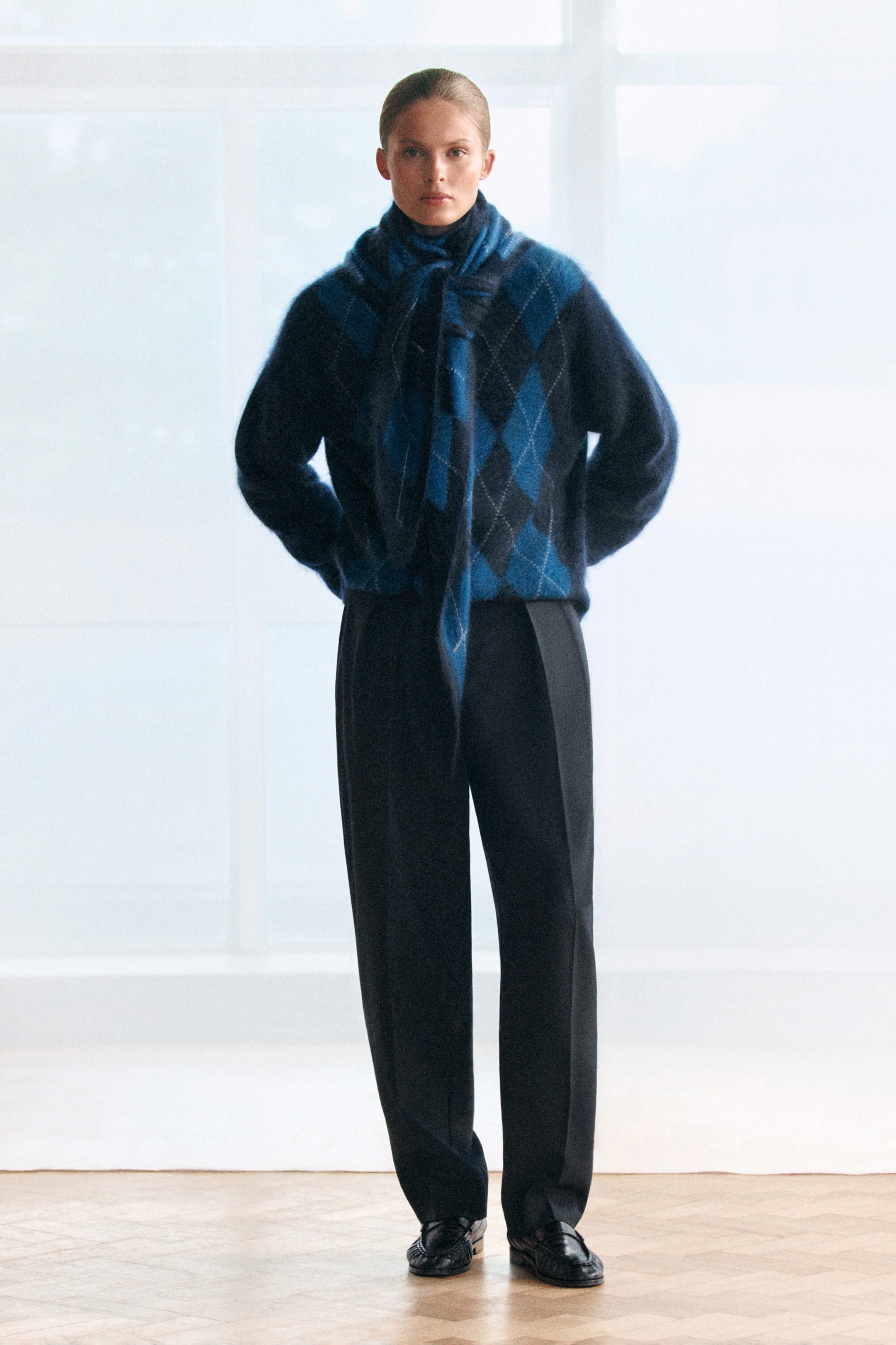 A model is wearing a front-facing, long-sleeved brushed cashmere jumper featuring a bold blue and black argyle pattern. The jumper is styled with a matching argyle knit scarf wrapped around the neck and paired with dark, wide-leg trousers. A model is wearing a front-facing, long-sleeved brushed cashmere jumper featuring a bold blue and black argyle pattern. The jumper is styled with a matching argyle knit scarf wrapped around the neck and paired with dark, wide-leg trousers.