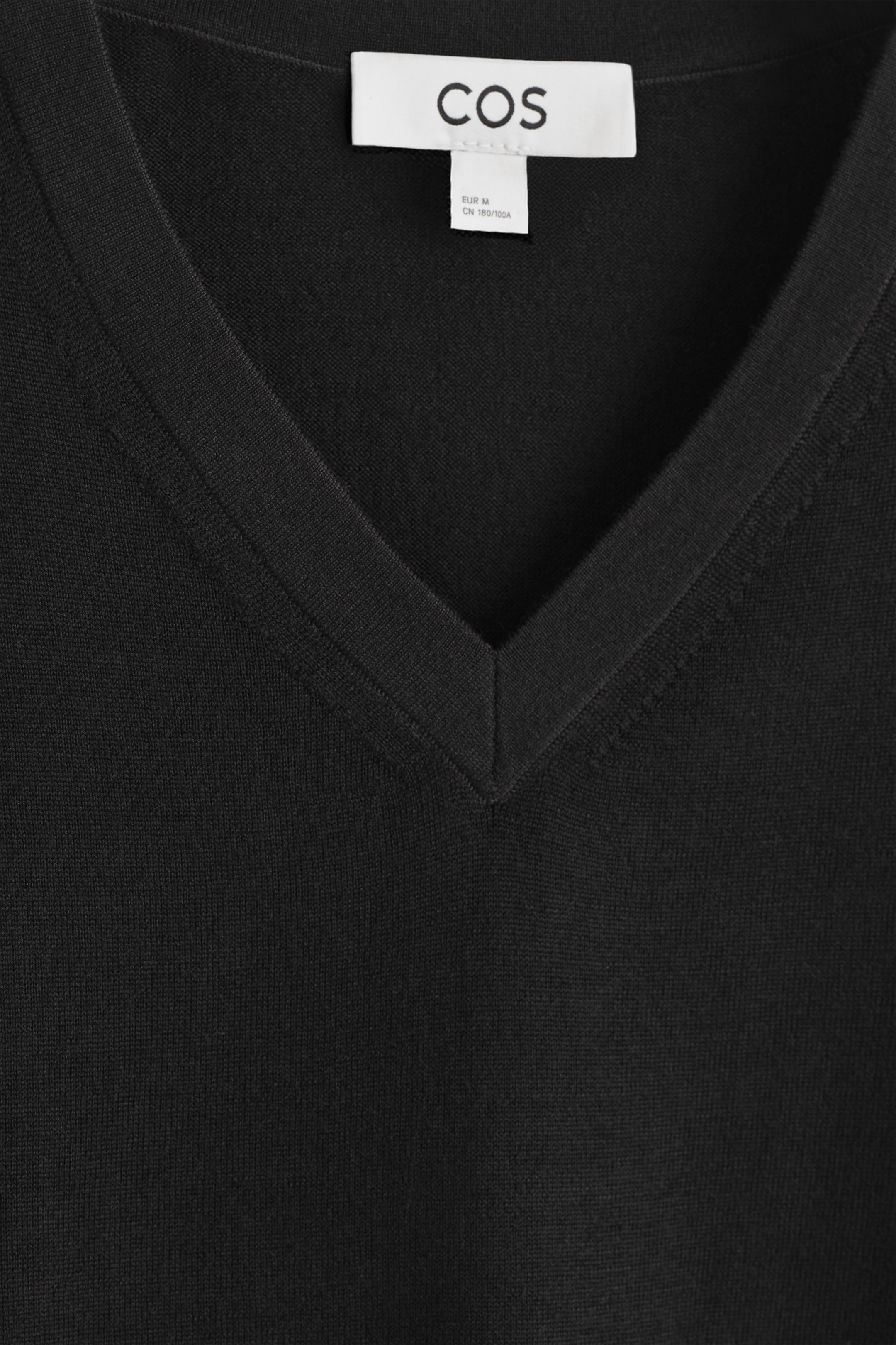 Visualizza immagine più grande: Un maglione nero con scollo a V è mostrato dalla PARTE ANTERIORE, caratterizzato da una trama a maglia fine e uno scollo a V stretto e rinforzato. Un'etichetta bianca del marchio 'COS' è visibile nella parte superiore dello scollo.