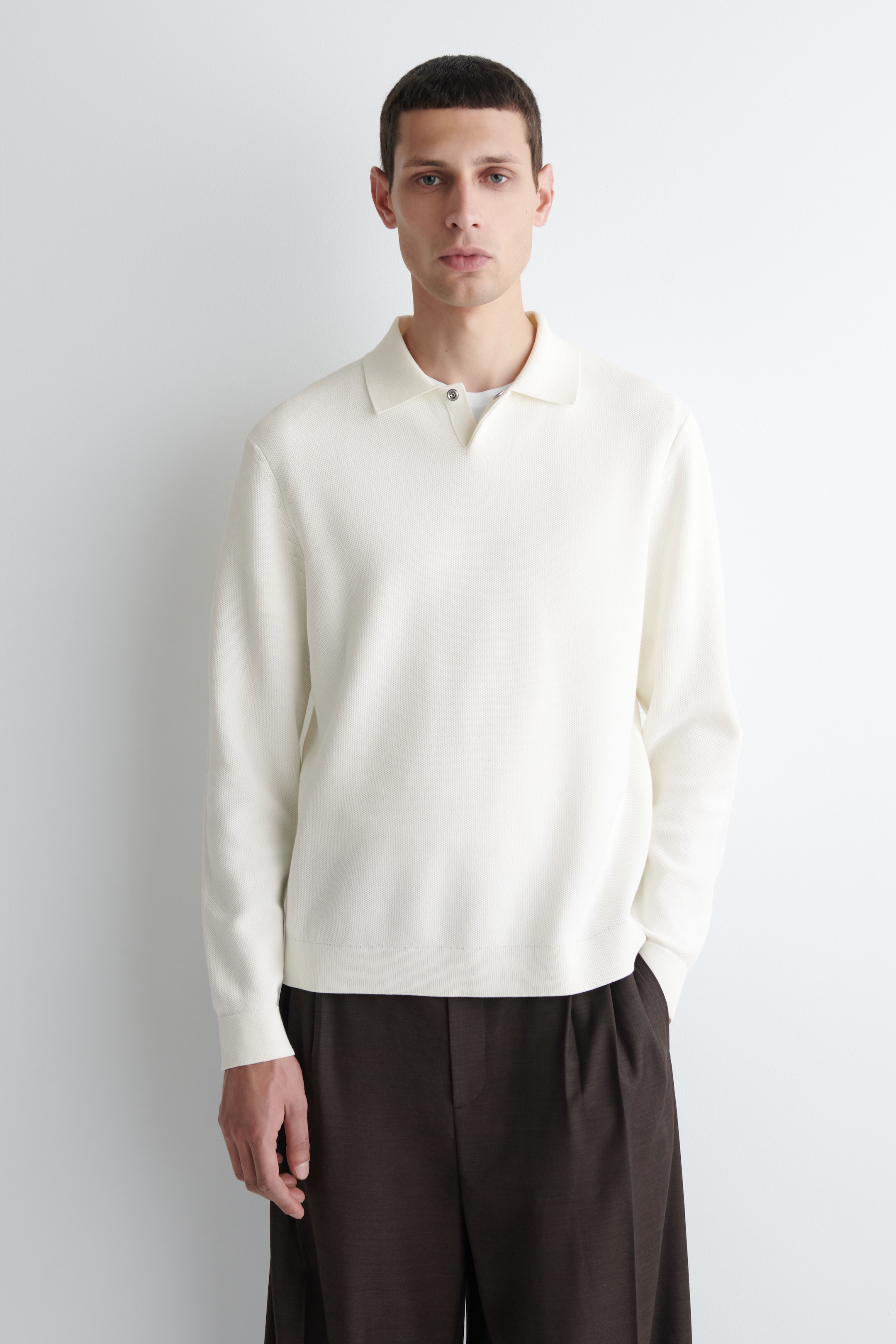 View larger image: A cream honeycomb-knit polo shirt is being shown from the front, featuring long sleeves with ribbed cuffs, a ribbed hem, a classic polo collar, and a notched V-neck opening secured by a single snap button, with a white undershirt visible beneath.