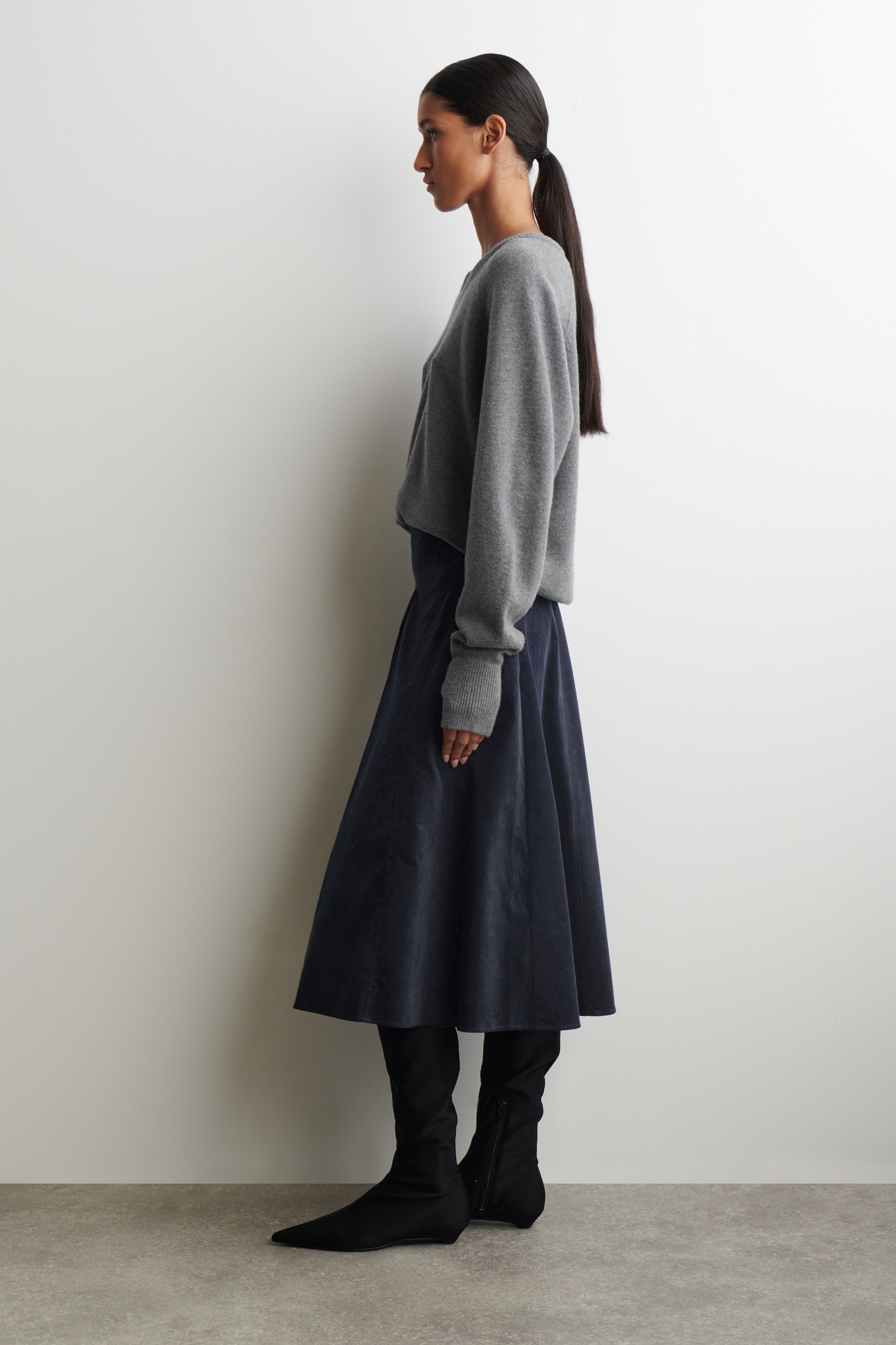 Agrandir l'image: Une jupe midi bleu marine foncé est présentée du côté gauche, montrant sa texture tactile en coton-corduroy avec des côtes apparentes. Elle présente une silhouette en ligne A avec un profond pli creux à l'avant, et s'évase depuis la taille tout en tombant sous les genoux, où elle rencontre des bottes noires à hauteur du genou.