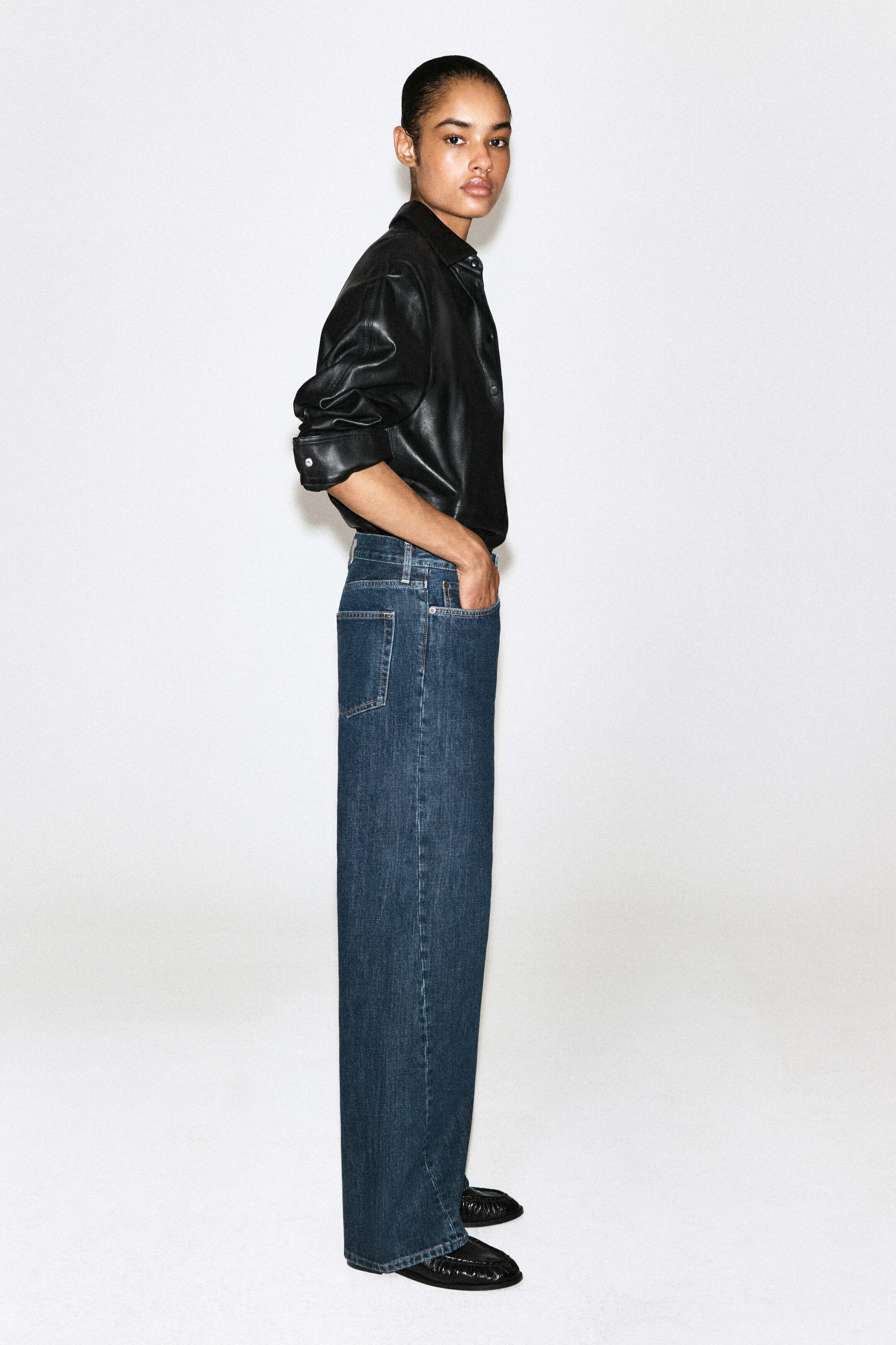 These wide-leg blue jeans in a medium wash are shown in a right-facing side profile, highlighting their relaxed straight-leg silhouette and classic five-pocket construction. The jeans are styled with a tucked-in black leather button-up shirt and black loafers. These wide-leg blue jeans in a medium wash are shown in a right-facing side profile, highlighting their relaxed straight-leg silhouette and classic five-pocket construction. The jeans are styled with a tucked-in black leather button-up shirt and black loafers.