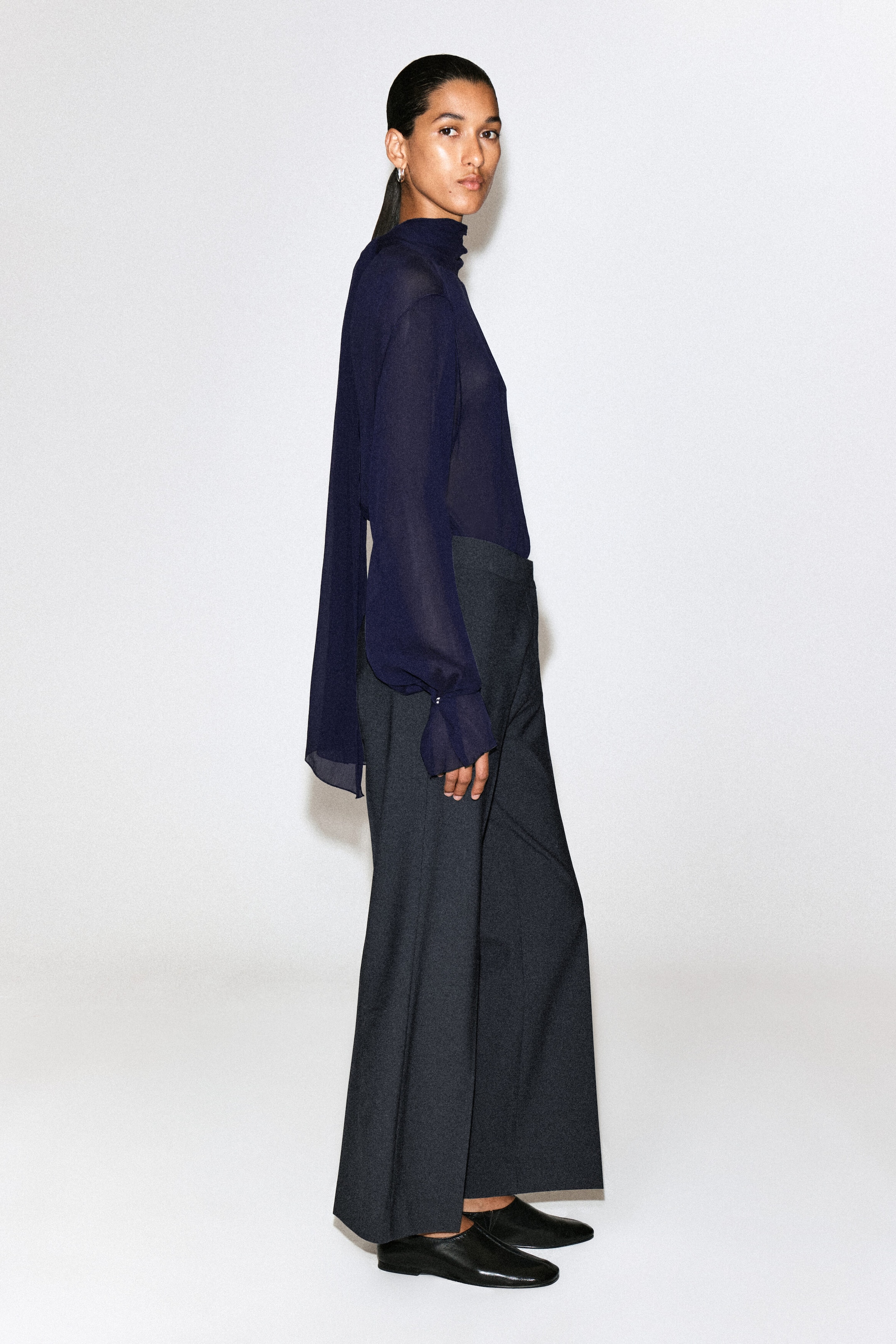 Twill cropped flared pants in dark grey are draping in a structured, wide-leg silhouette, presented from a side profile facing right to highlight their high-waisted fit and ankle-length hem. The pants are being styled with a tucked-in sheer navy blue blouse and black flat leather shoes to emphasize their clean, tailored design.