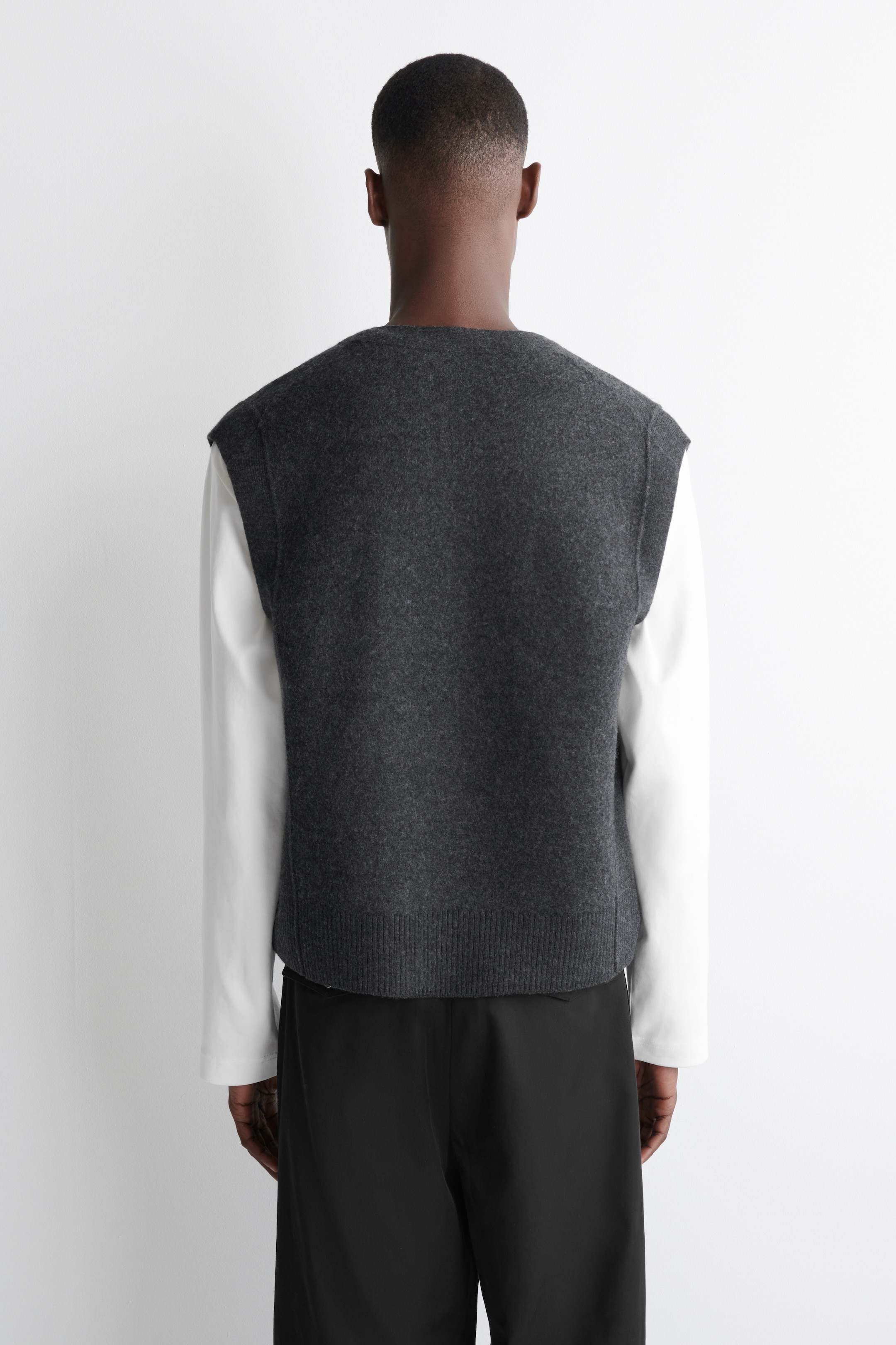 View larger image: A dark grey knit vest is being presented from the rear. It is layered over a white long-sleeved shirt and is showcasing a rounded neckline and a ribbed hem, resting at the waist of black trousers.