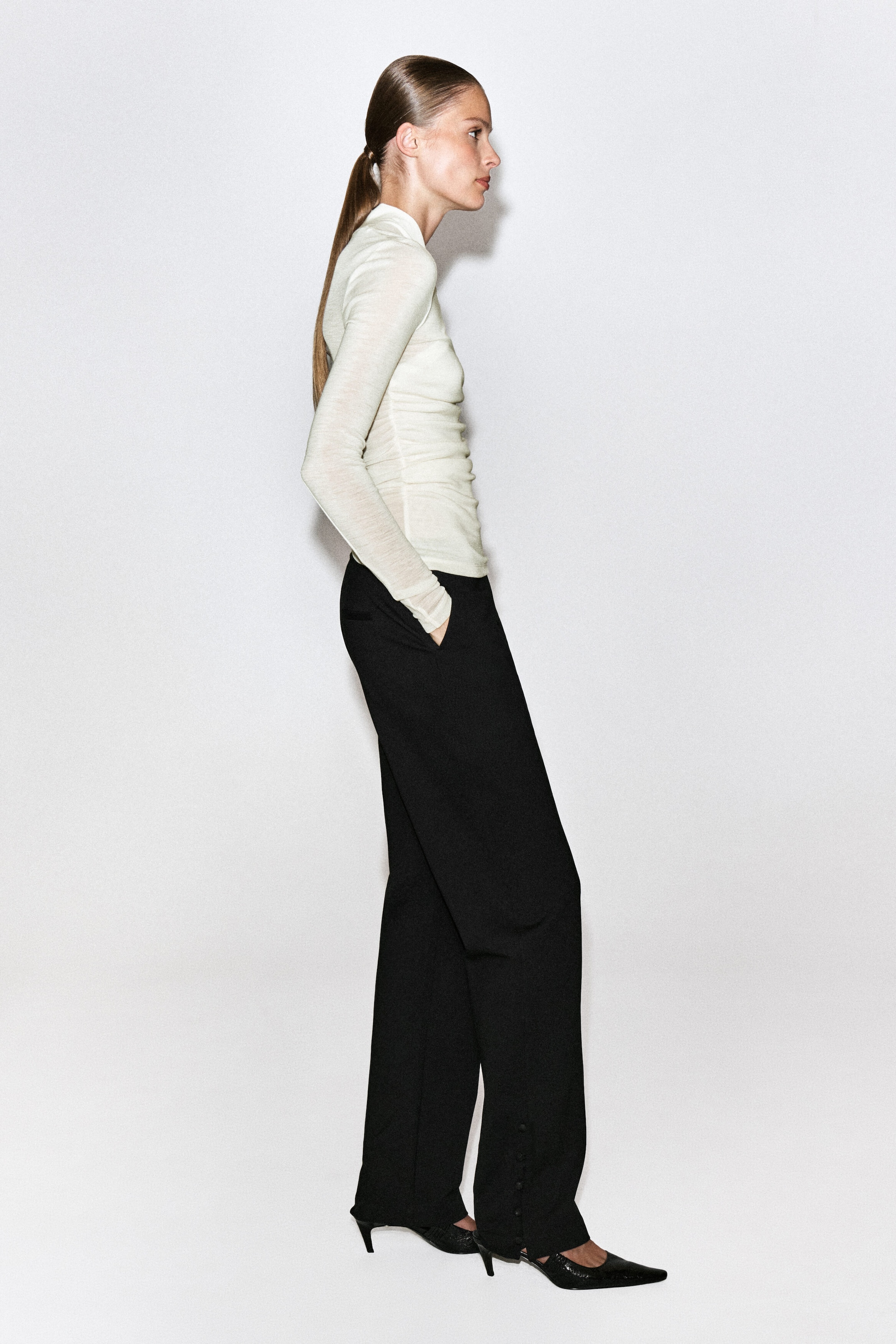 A cream-coloured, slim-fit, long-sleeved top featuring a high collar and gathered detailing is being worn by the model. The model is styling the top with black wide-leg trousers and black pointed-toe heels. A cream-coloured, slim-fit, long-sleeved top featuring a high collar and gathered detailing is being worn by the model. The model is styling the top with black wide-leg trousers and black pointed-toe heels.