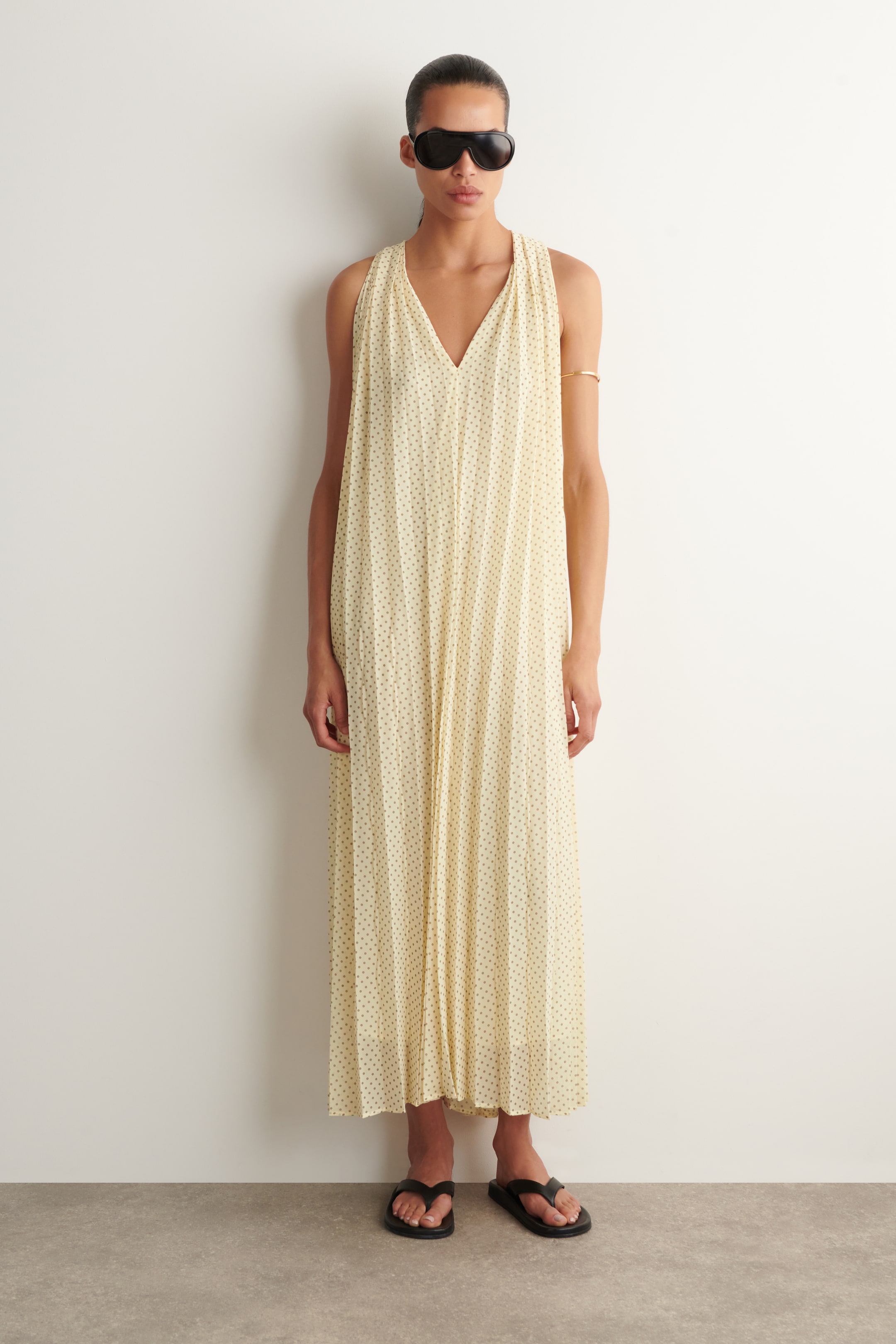 View larger image: A cream-coloured, sleeveless jumpsuit featuring a V-neckline and a subtle dark polka dot pattern, with vertical pleats creating a loose, flowing silhouette.