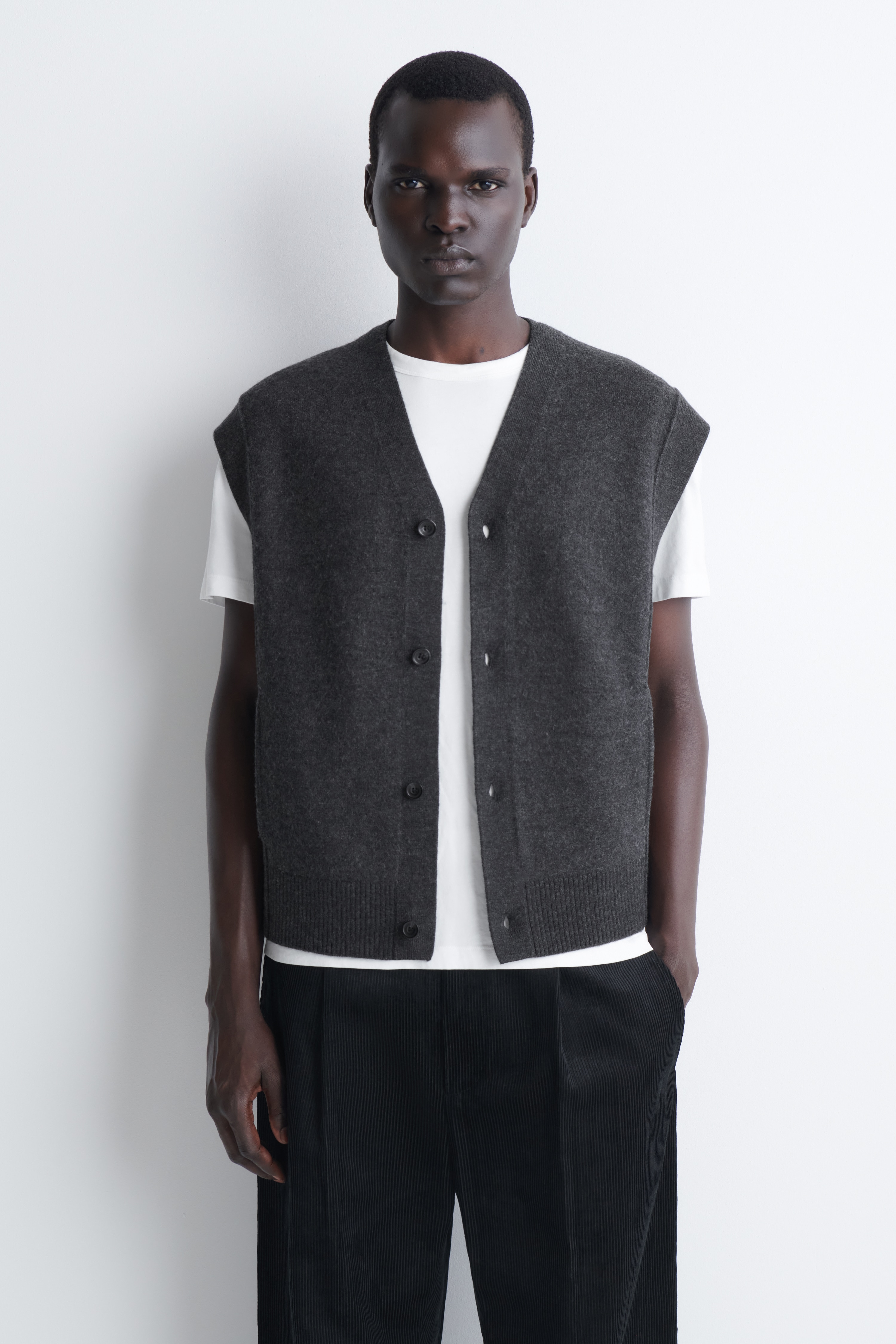 The buttoned boiled-wool waistcoat in dark grey is shown front-on, featuring a deep V-neckline and a four-button front closure. The sleeveless design incorporates subtle side welt pockets and a ribbed hem.