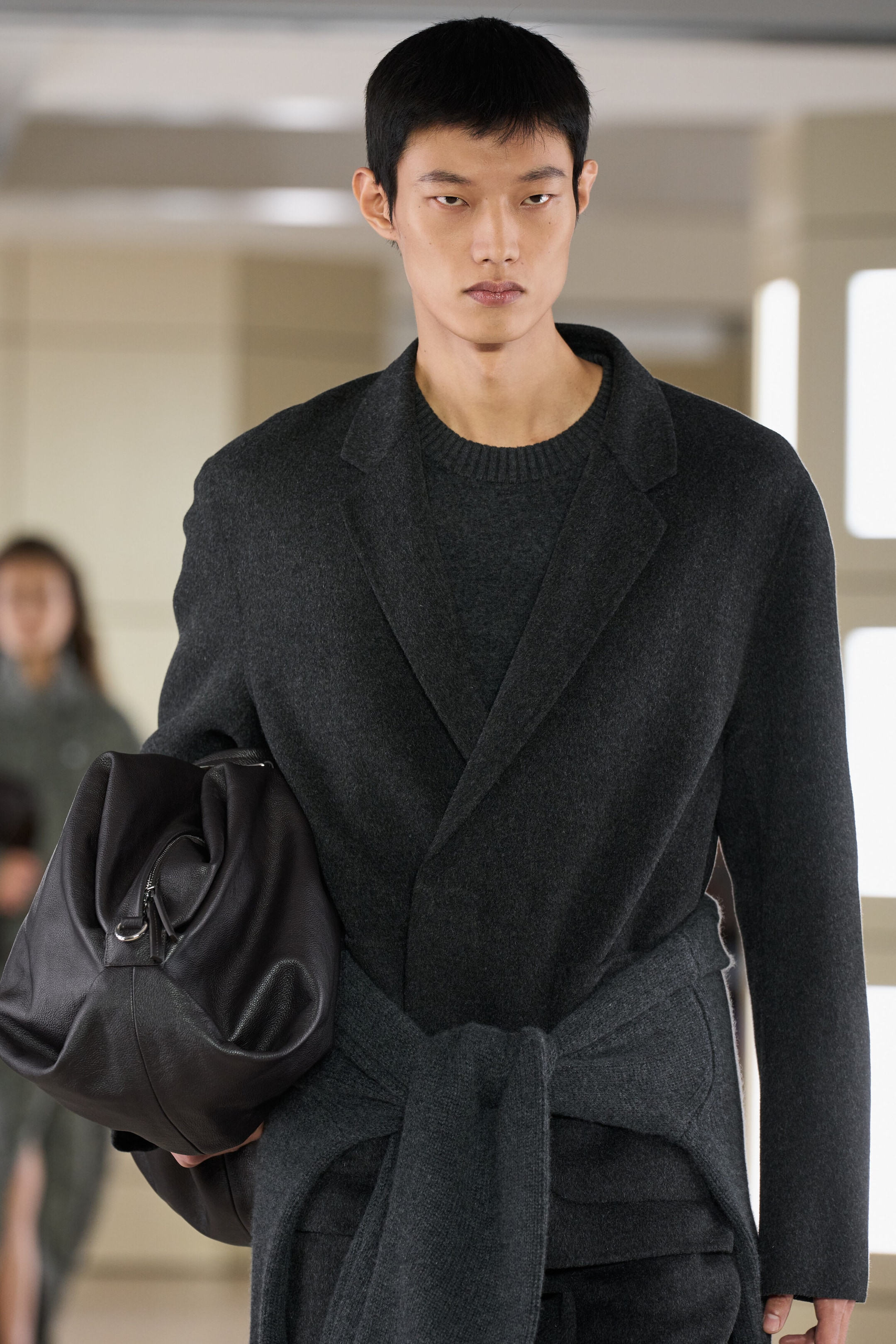 Facing front, a model is wearing a relaxed-fit, dark grey wool-blend blazer with notch lapels, layered over a matching crew-neck knit jumper. They are carrying a slouchy, black leather duffle bag with a silver zip closure tucked under one arm.