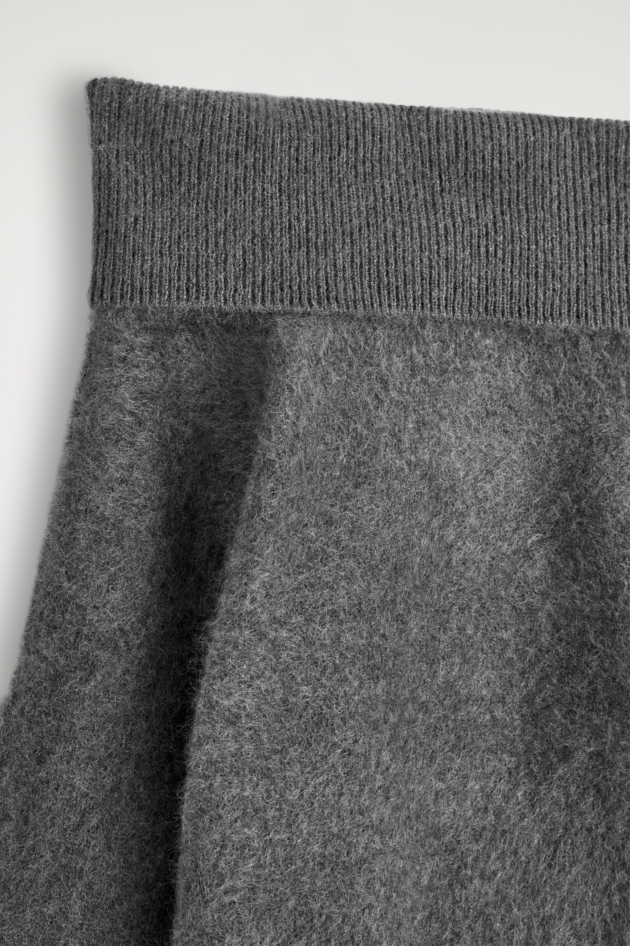 Ingrandisci l'immagine: Un maglione grigio in maglia presenta una consistenza spazzolata e pelosa sul corpo principale. Il bordo superiore mostra un'ampia fascia a coste ripiegata.