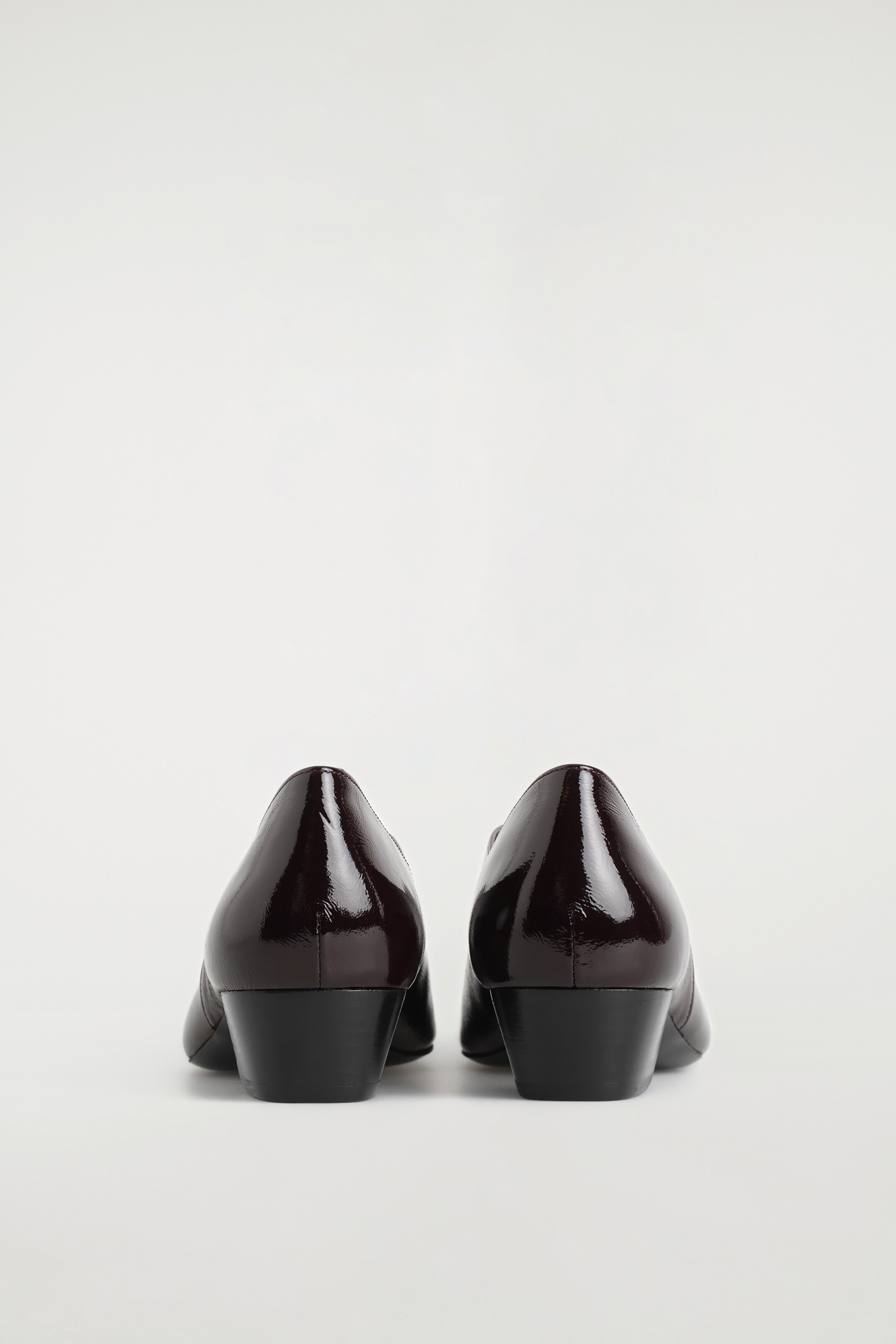 Agrandir l'image: Deux ballerines modernes bordeaux sont debout vues de dos. Elles réfléchissent la lumière grâce à leur cuir verni, présentent une silhouette angulaire et reposent sur de petits talons empilés.