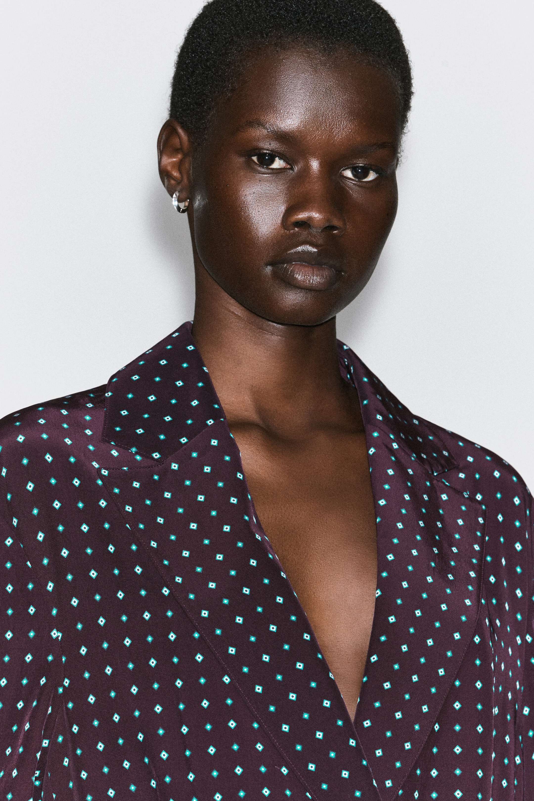 The diamond-print double-breasted blazer is shown from the front, featuring a deep plum fabric patterned with small teal and white geometric diamonds. The blazer features classic notch lapels and a deep V-neckline, highlighting its smooth, lustrous material. The diamond-print double-breasted blazer is shown from the front, featuring a deep plum fabric patterned with small teal and white geometric diamonds. The blazer features classic notch lapels and a deep V-neckline, highlighting its smooth, lustrous material.