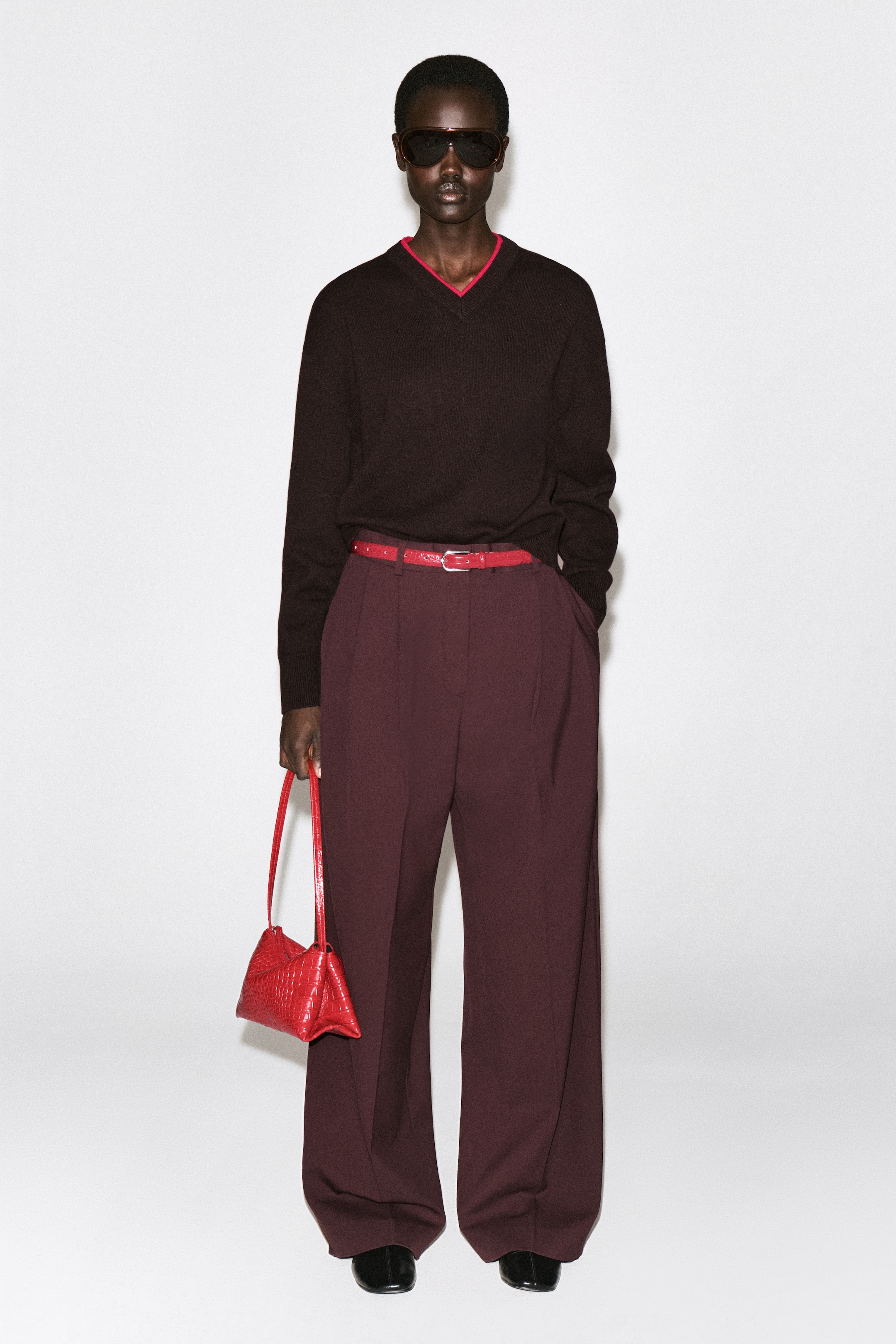 Größeres Bild anzeigen: Die rote Mini-Pavilion-Shoulder-Bag aus Leder hängt an der Hand eines Models in frontaler Ansicht und präsentiert ihre strukturierte, dreieckige Silhouette. Die Tasche zeigt eine strukturierte Oberfläche mit Krokodil-Effekt und einen schmalen, farblich abgestimmten Schulterriemen.