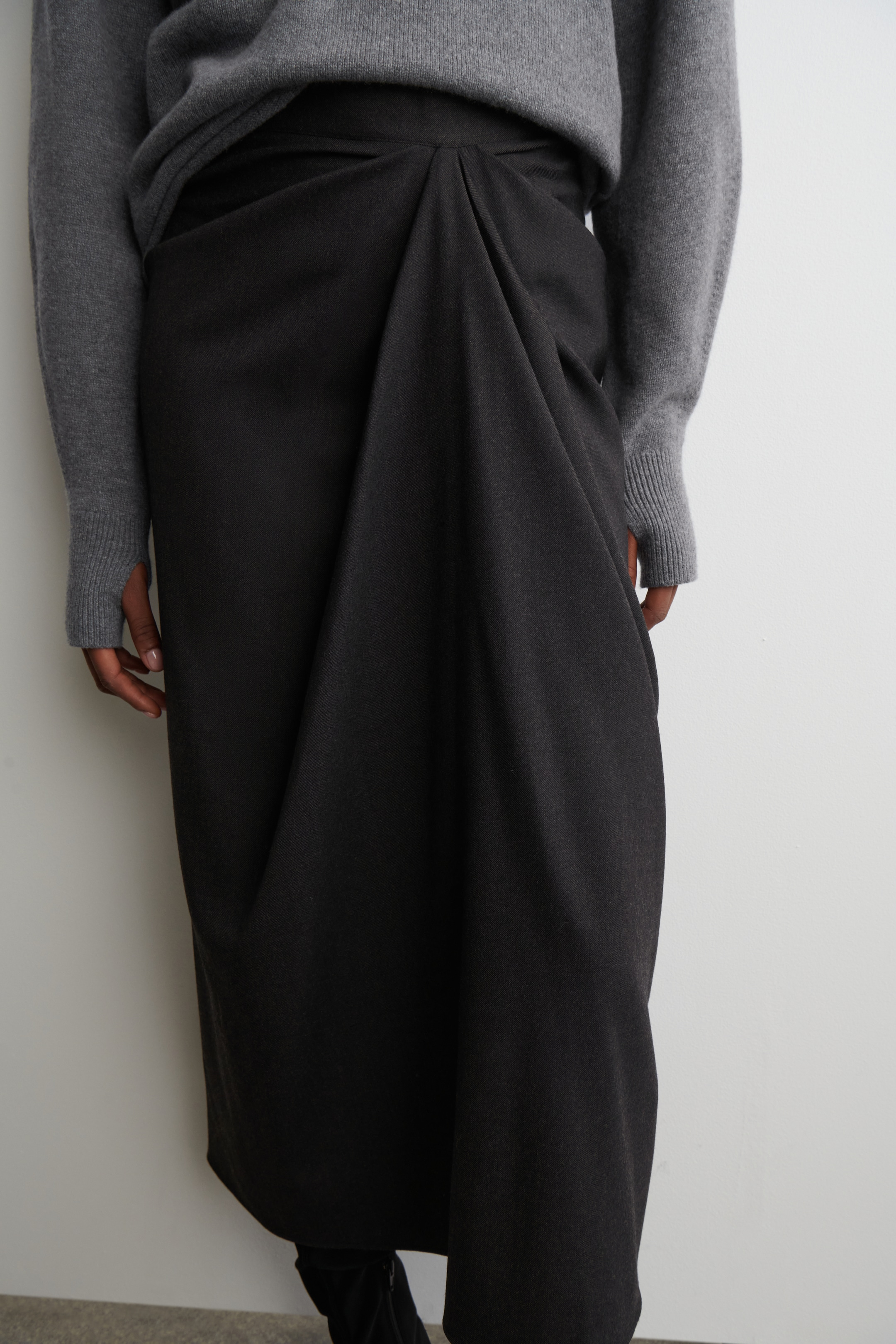 Nagyobb kép megtekintése: A sötétszürke DRAPED WOOL-BLEND MIDI SZOKNYÁT a modell viseli, amelynek jellegzetes elülső drapériája van. A világosszürke kötött pulóver, amelyen látható bordázott mandzsetták és hüvelykujjlyukak vannak, szintén a szoknya fölött van viselve.