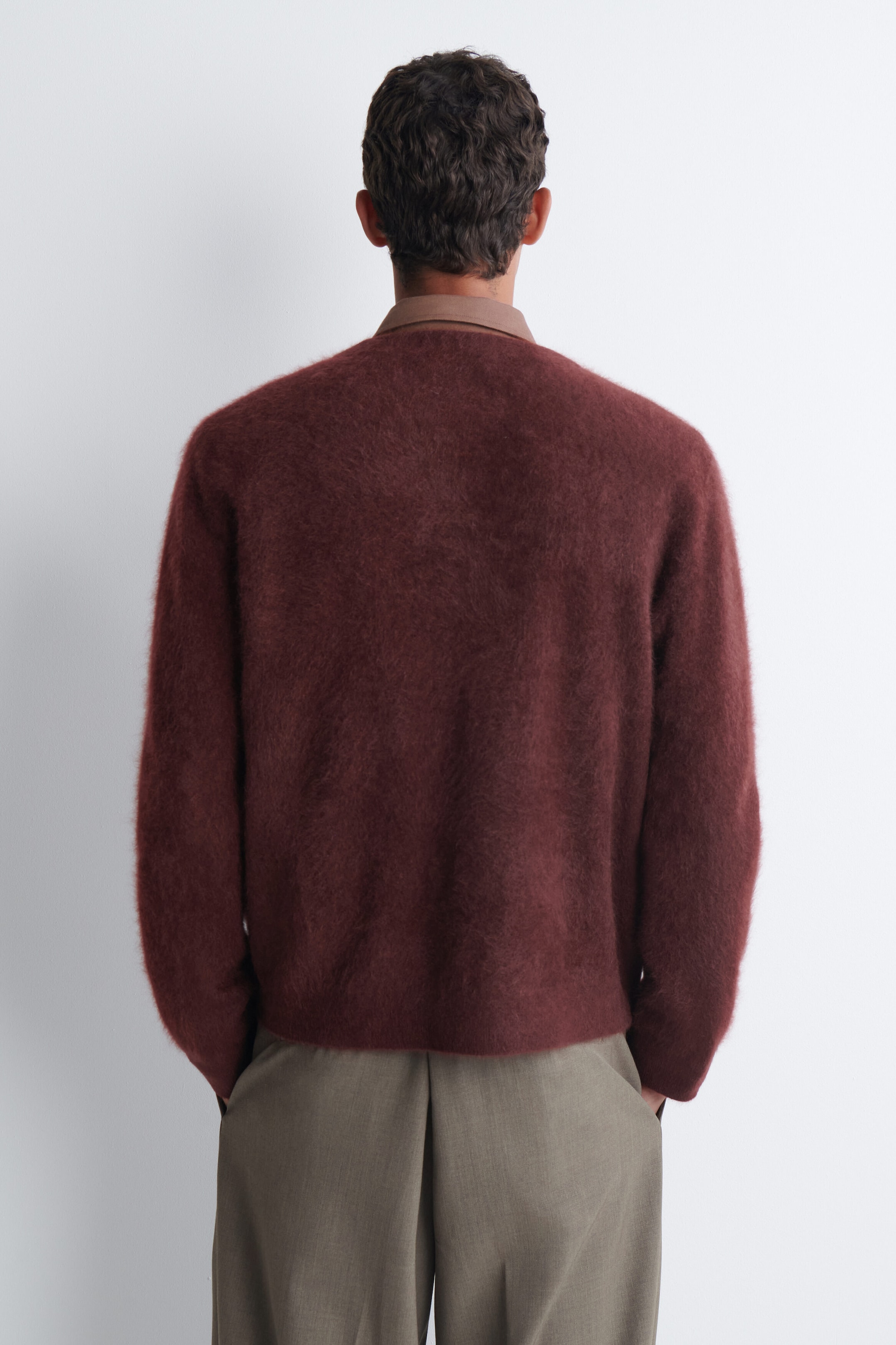 A burgundy brushed-cashmere cardigan with a fuzzy texture is worn by the model, shown from the back. The cardigan is layered over a brown collared shirt and paired with olive-grey trousers. A burgundy brushed-cashmere cardigan with a fuzzy texture is worn by the model, shown from the back. The cardigan is layered over a brown collared shirt and paired with olive-grey trousers.