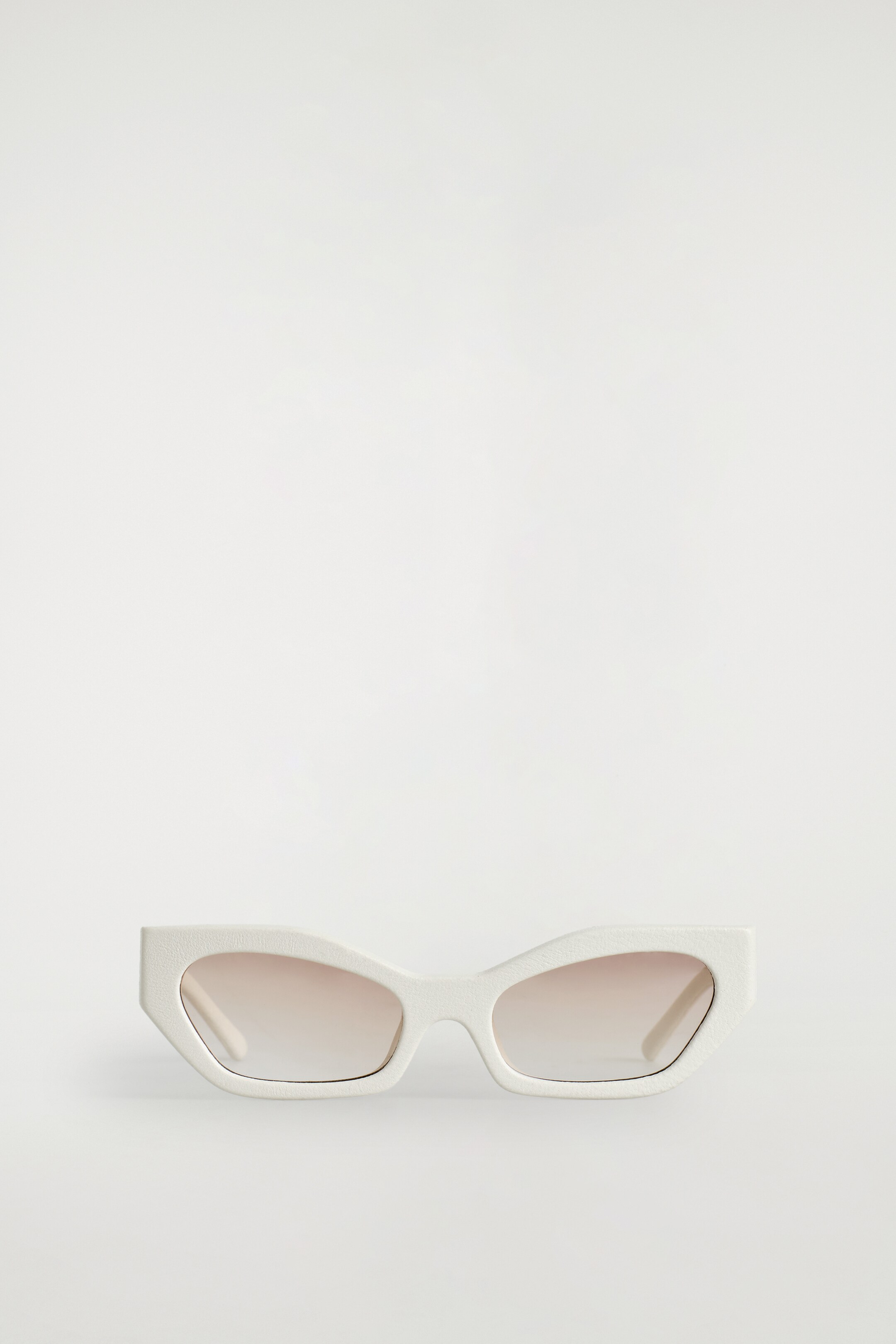 View larger image: White angular cat-eye sunglasses are being presented from a front angle, featuring a subtly textured frame and gradient brown-to-clear lenses.