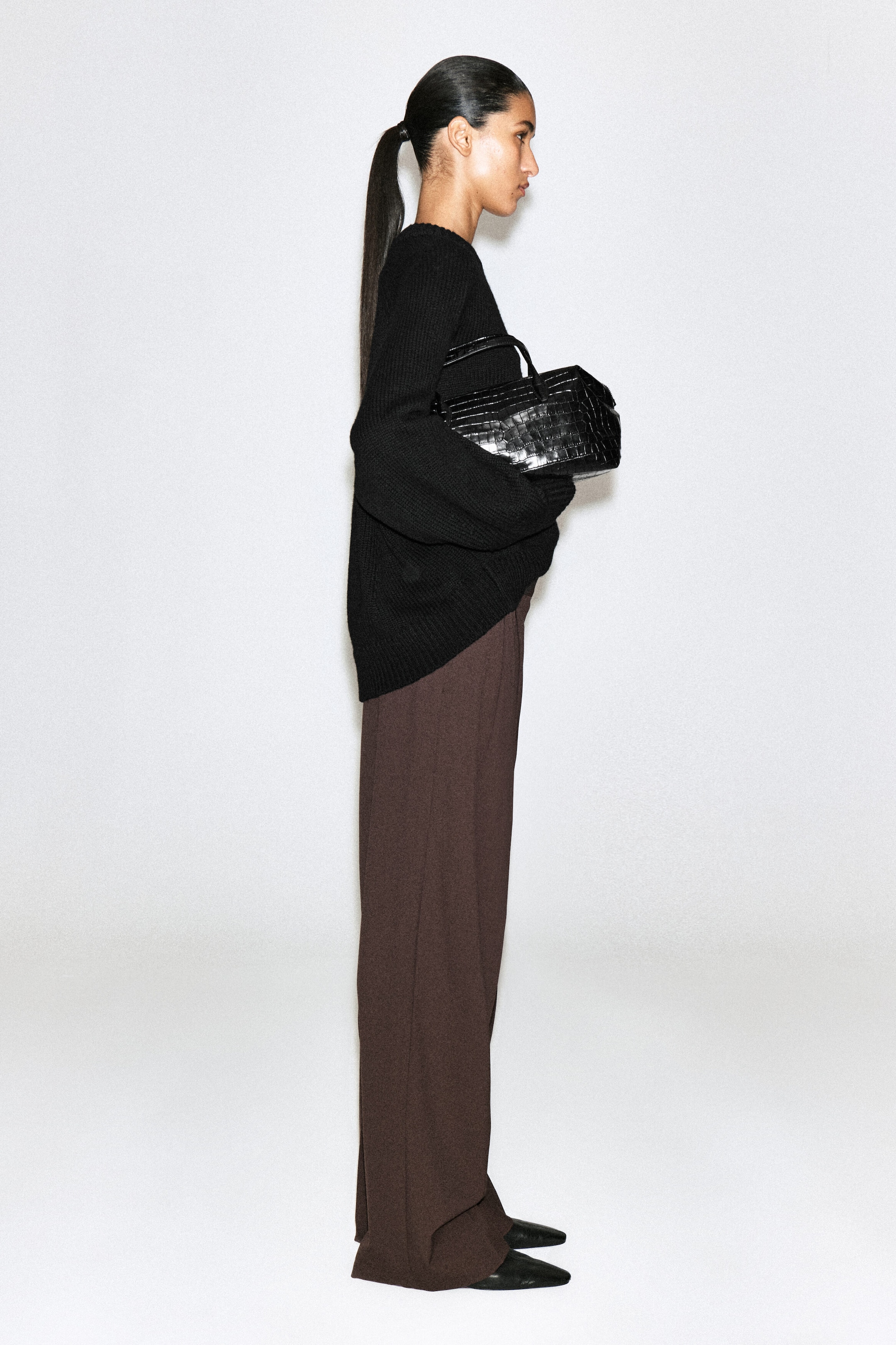A model facing right is wearing a black ribbed-knit crew-neck sweater and brown wide-leg trousers. The model is holding a black, structured, crocodile-embossed handbag with top handles, while black pointed-toe shoes peek out from beneath the trousers.